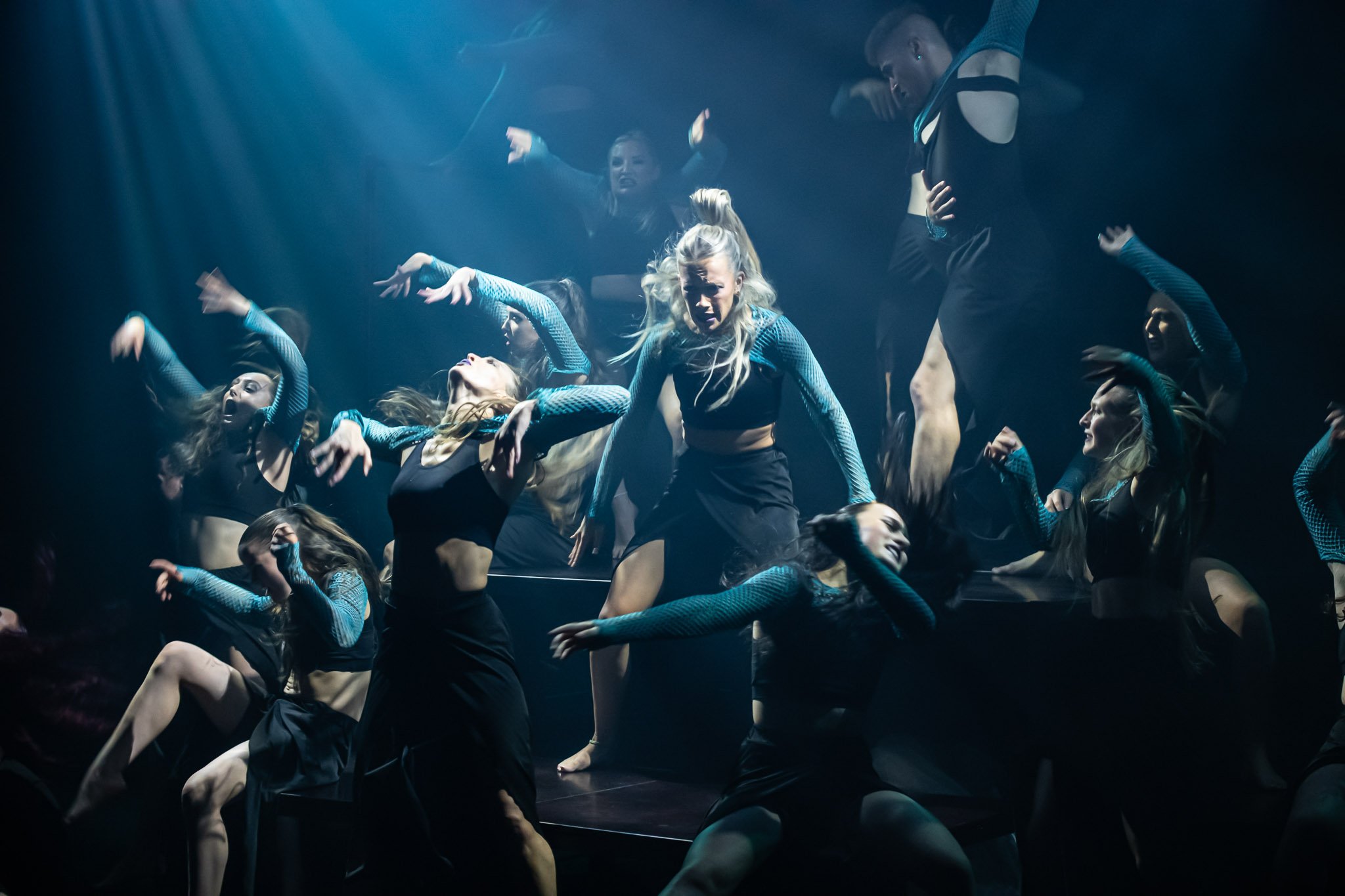 Group of dancers performing energetically with dramatic lighting on stage, wearing black outfits with patterned sleeves.