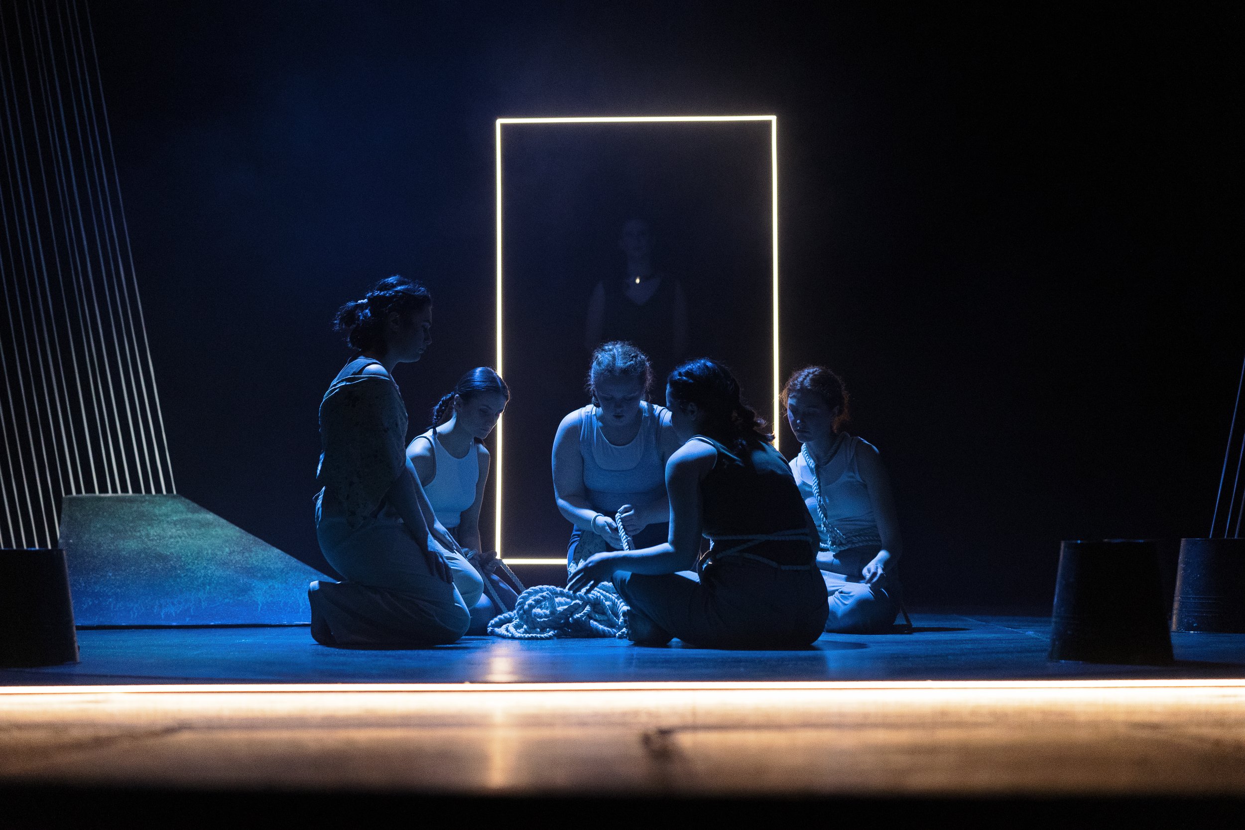 Group of women sitting on stage with rope, dark setting, glowing rectangular frame in background.