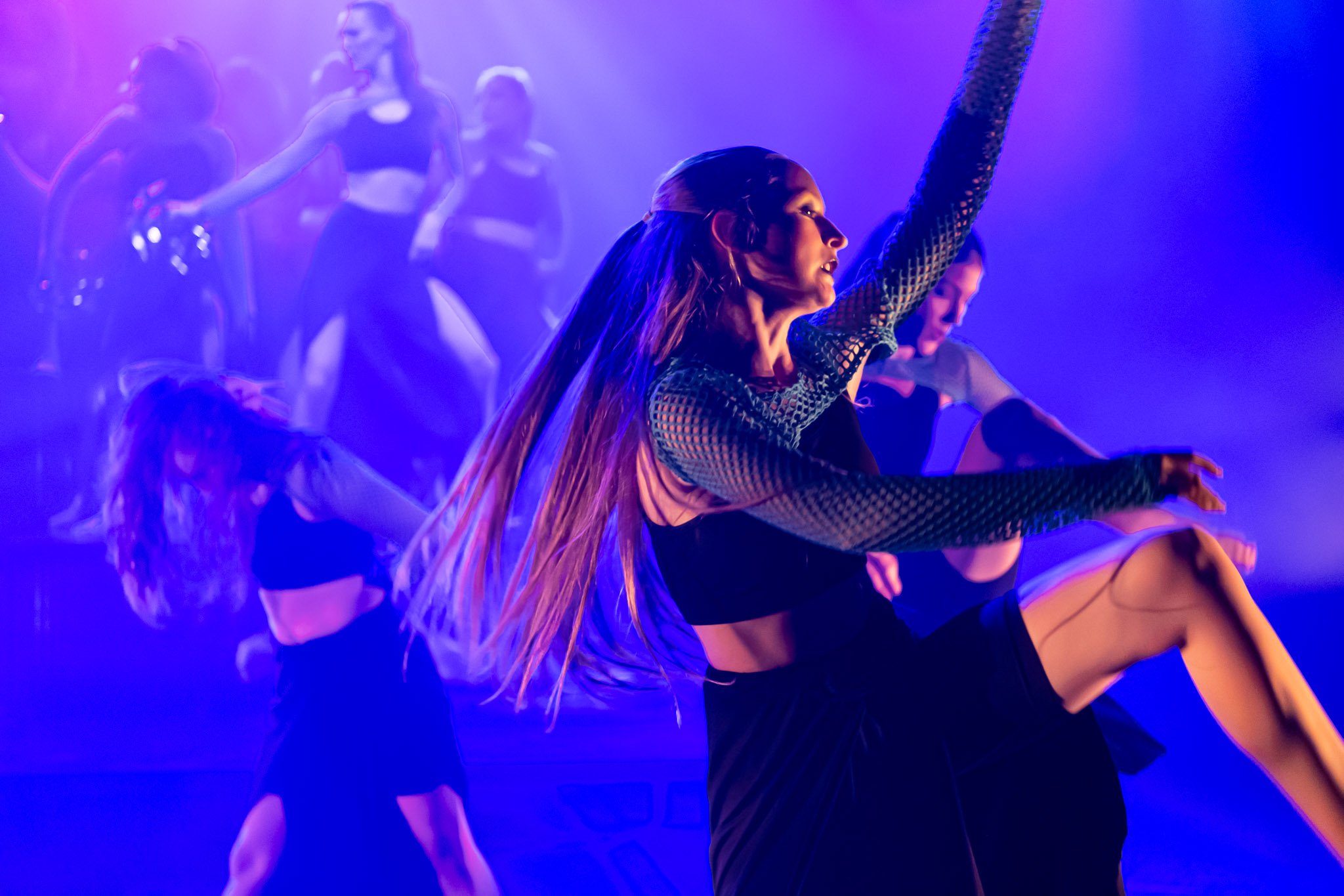 Group of female dancers performing on stage with colorful lighting, wearing black outfits.