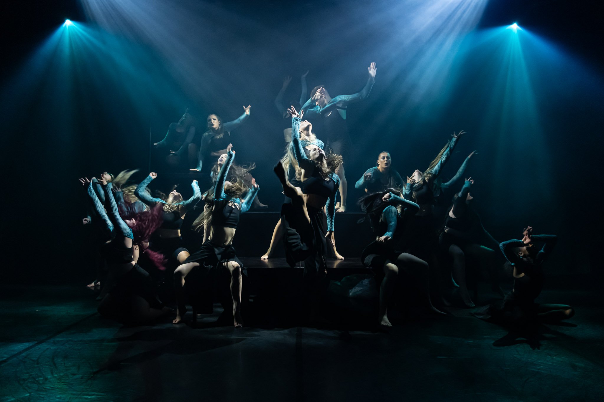 Group of dancers performing on stage under dramatic lighting, wearing dark costumes with blue accents, striking expressive poses.