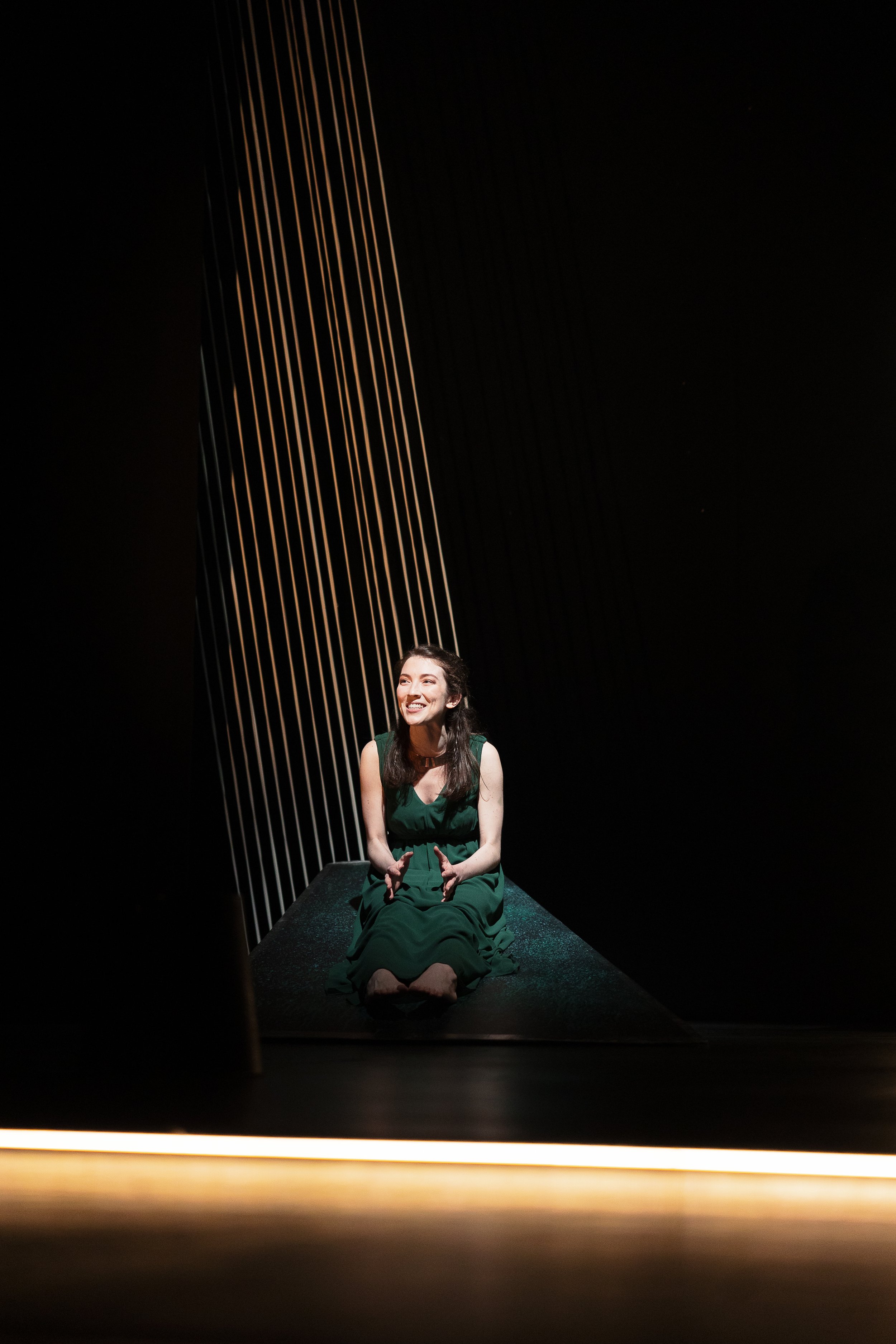 Woman in a green dress sitting on a dark stage with vertical light beams and smiling.