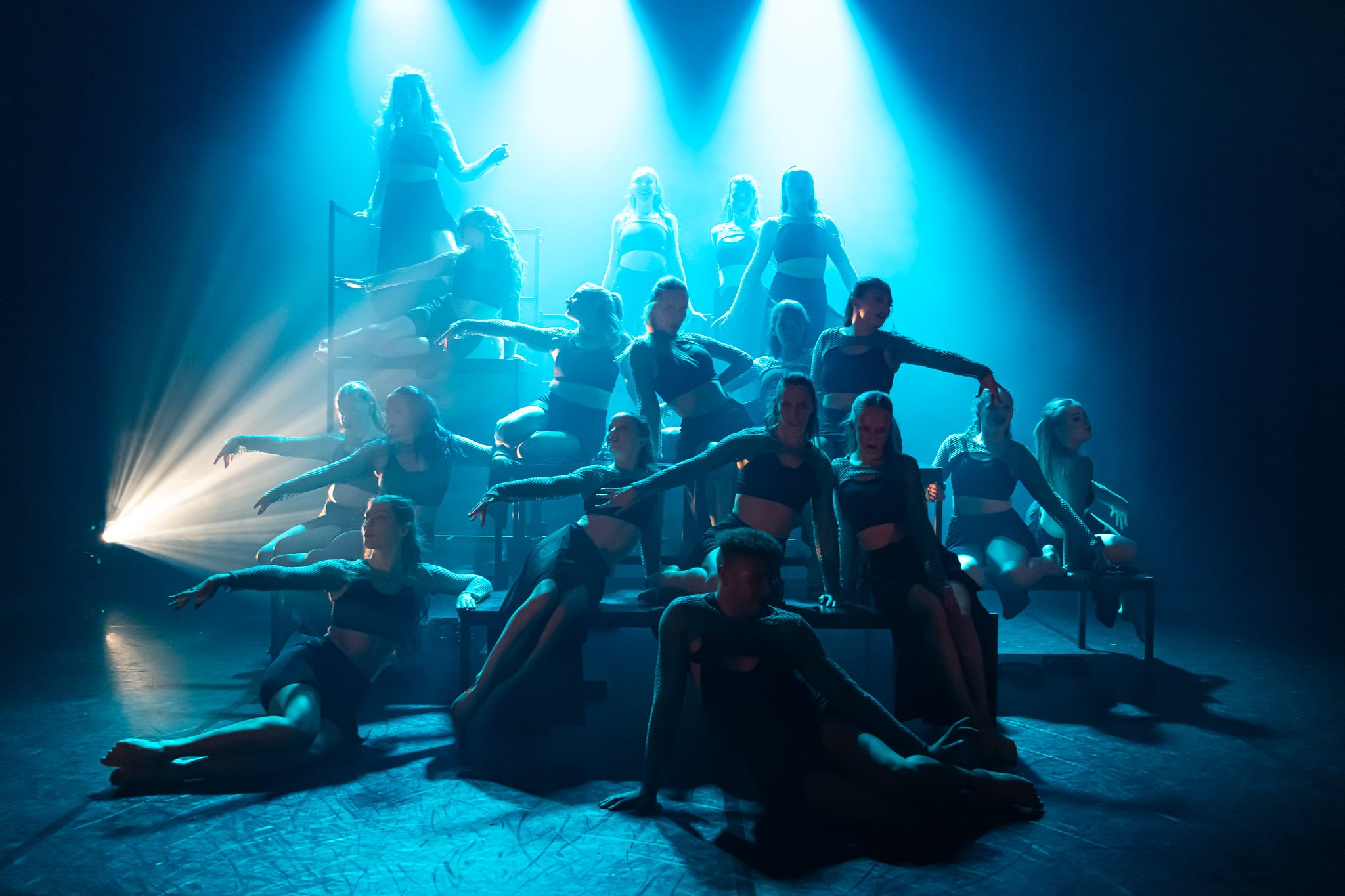 Group of dancers performing on a dimly lit stage with blue lighting and dramatic poses, wearing black costumes.