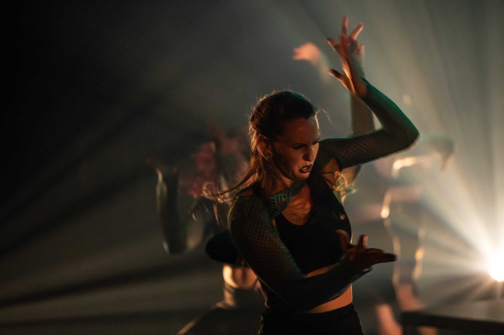 Female dancer performing energetically in dramatic lighting with others in the background.