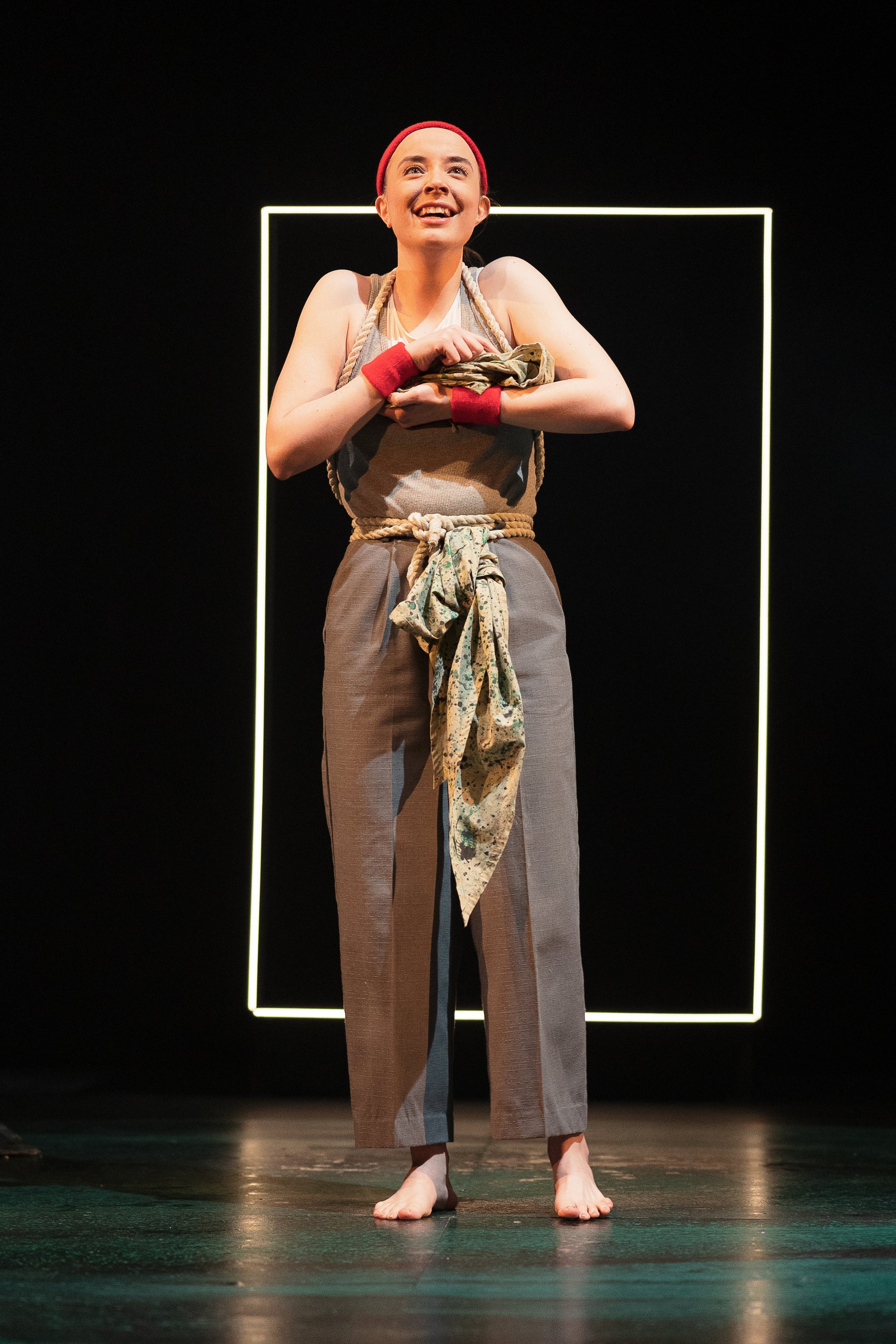 A person on stage wearing a red headband and wristbands, a gray sleeveless outfit, and a rope and fabric tied around the waist. They hold something close to their chest and are framed by a glowing outline in the background.