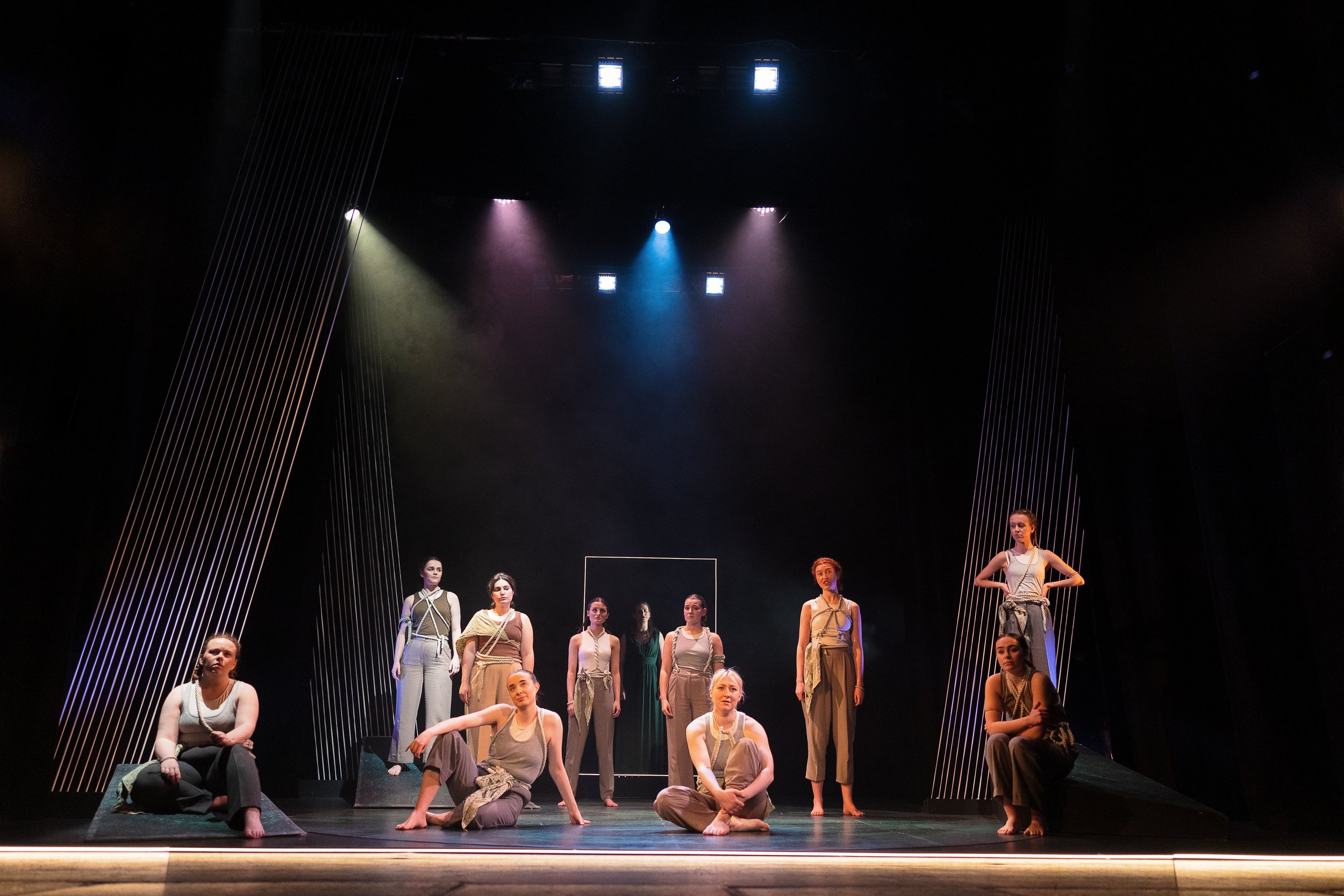 Group of performers on stage in modern dance or theater production, wearing neutral-colored costumes, with dramatic lighting and abstract set design.