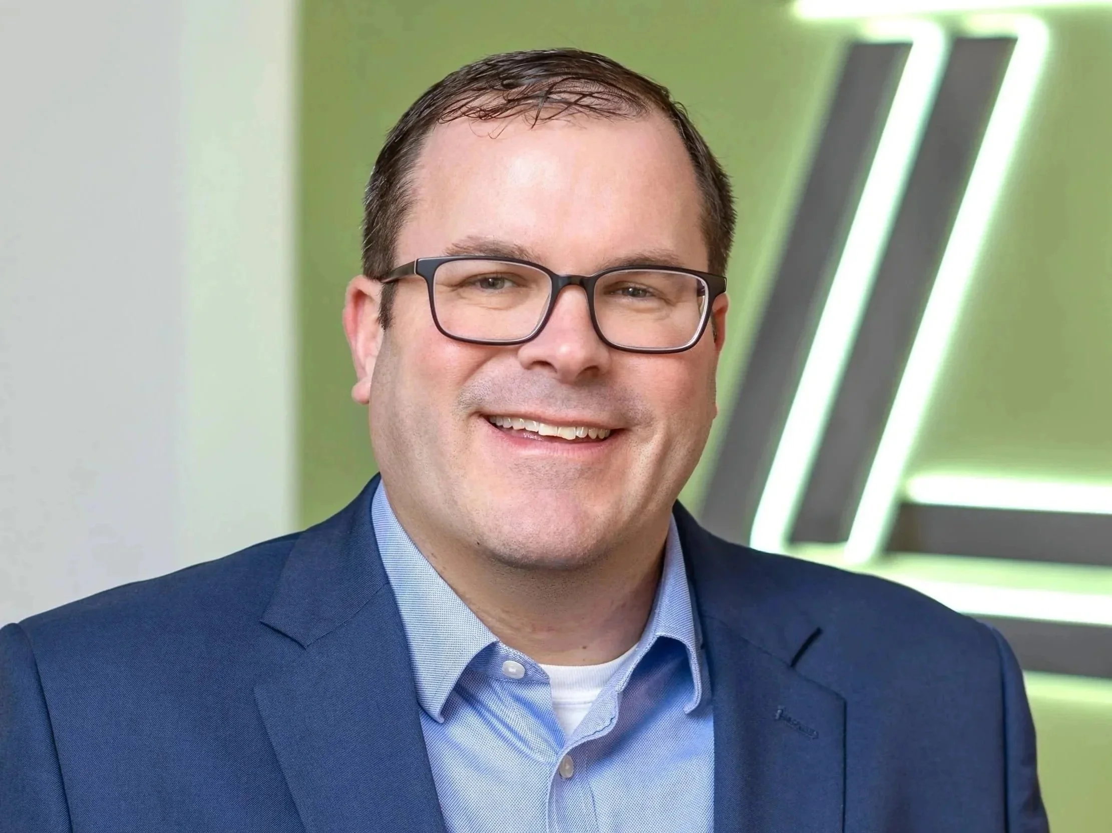 Headshot of a smiling man with glasses, wearing a blue suit and light blue shirt, in front of a green background with modern light fixtures.