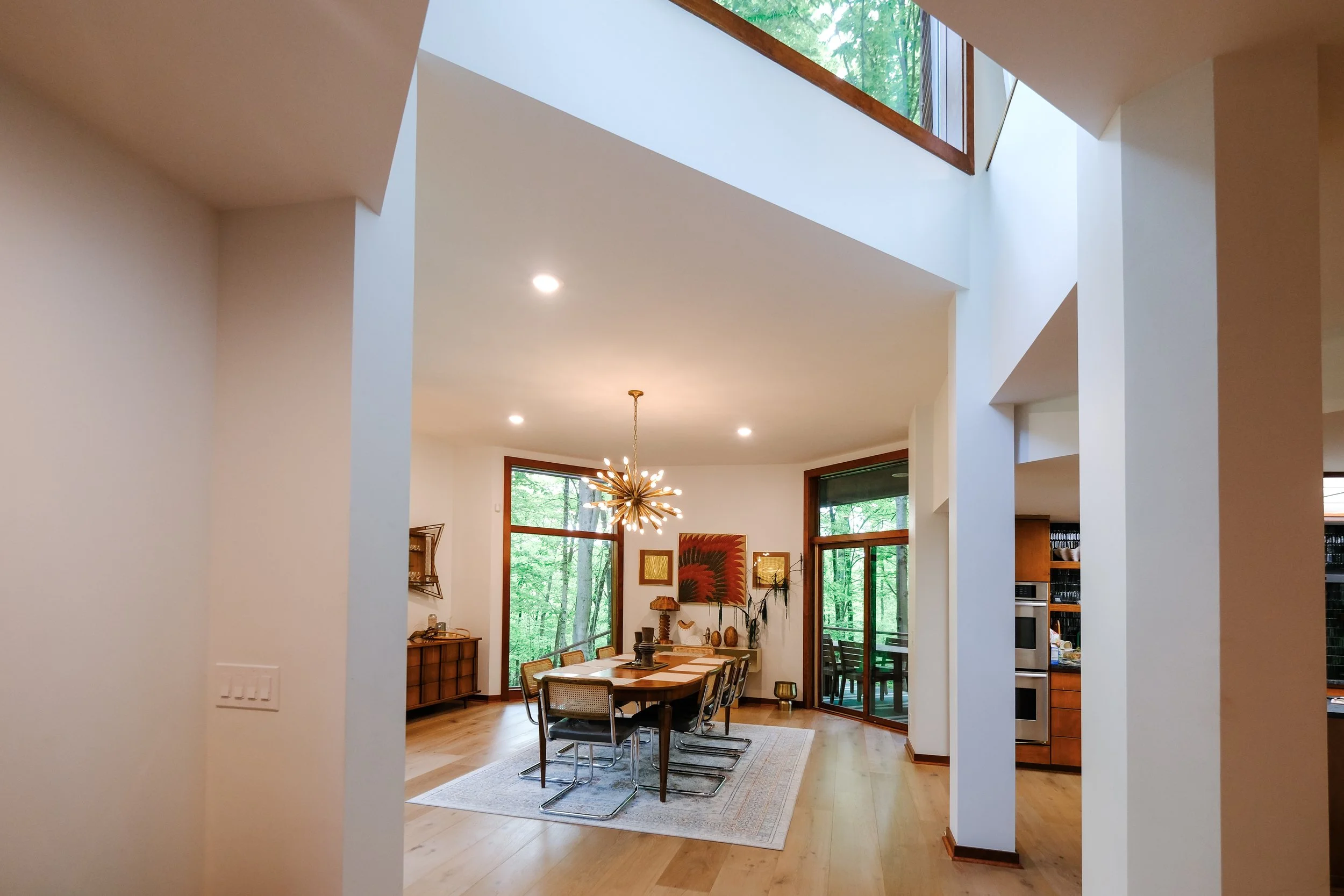 Modern dining room with large windows, a wooden dining table, six chairs, a chandelier, and artwork on the walls, with a view of a forest outside.