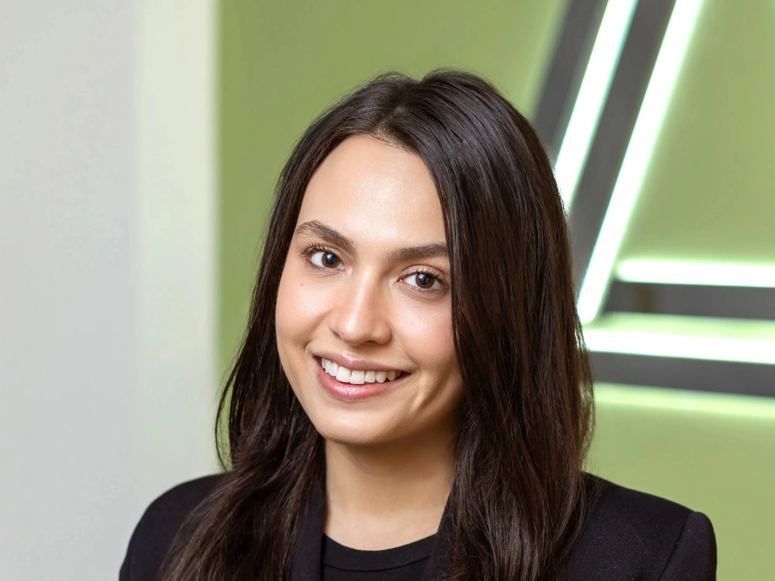 A young woman with long dark hair and a black jacket, smiling at the camera, with a green background and modern lighting.
