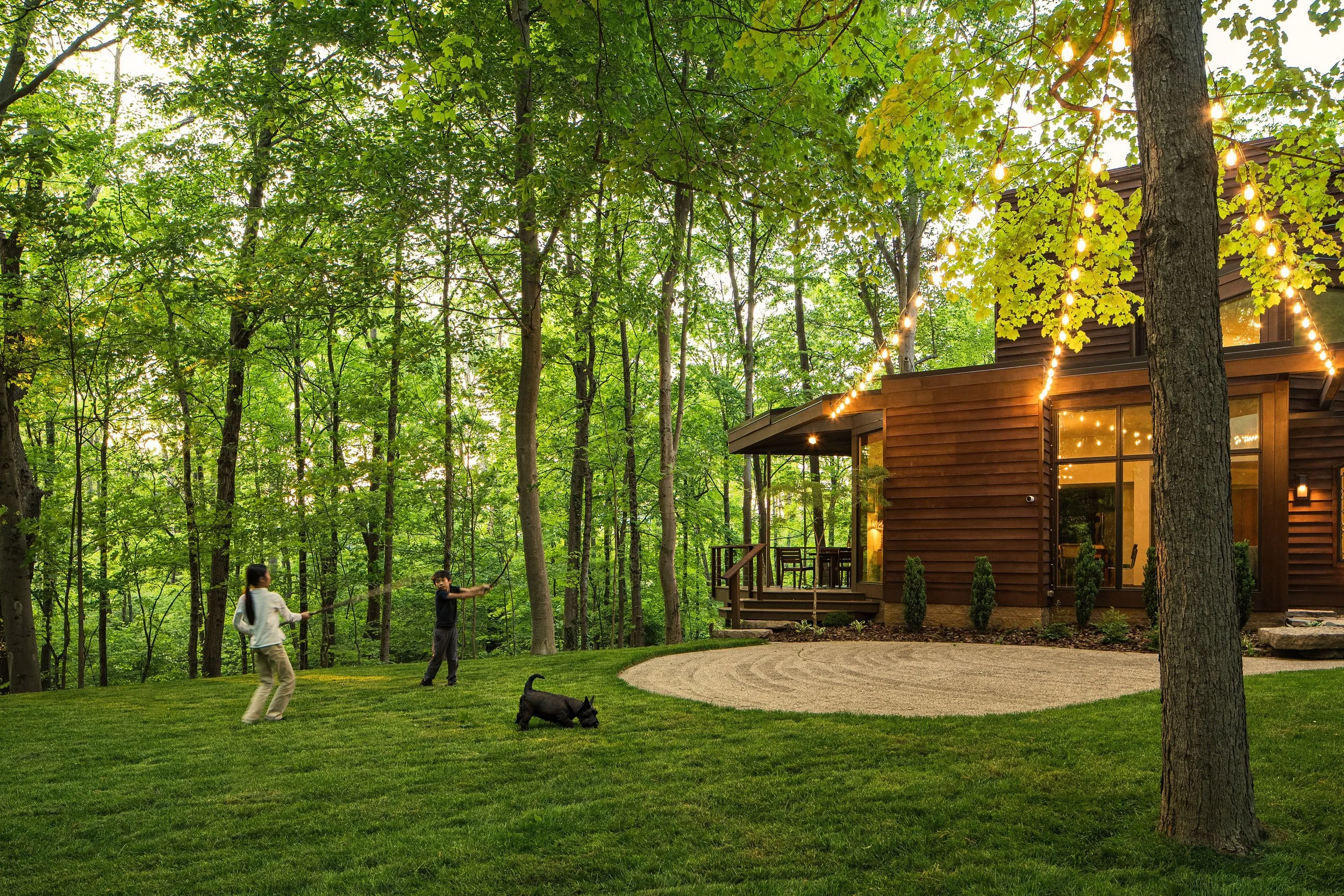 Children playing with a dog on a grassy backyard near a wooden house with string lights in a wooded area.