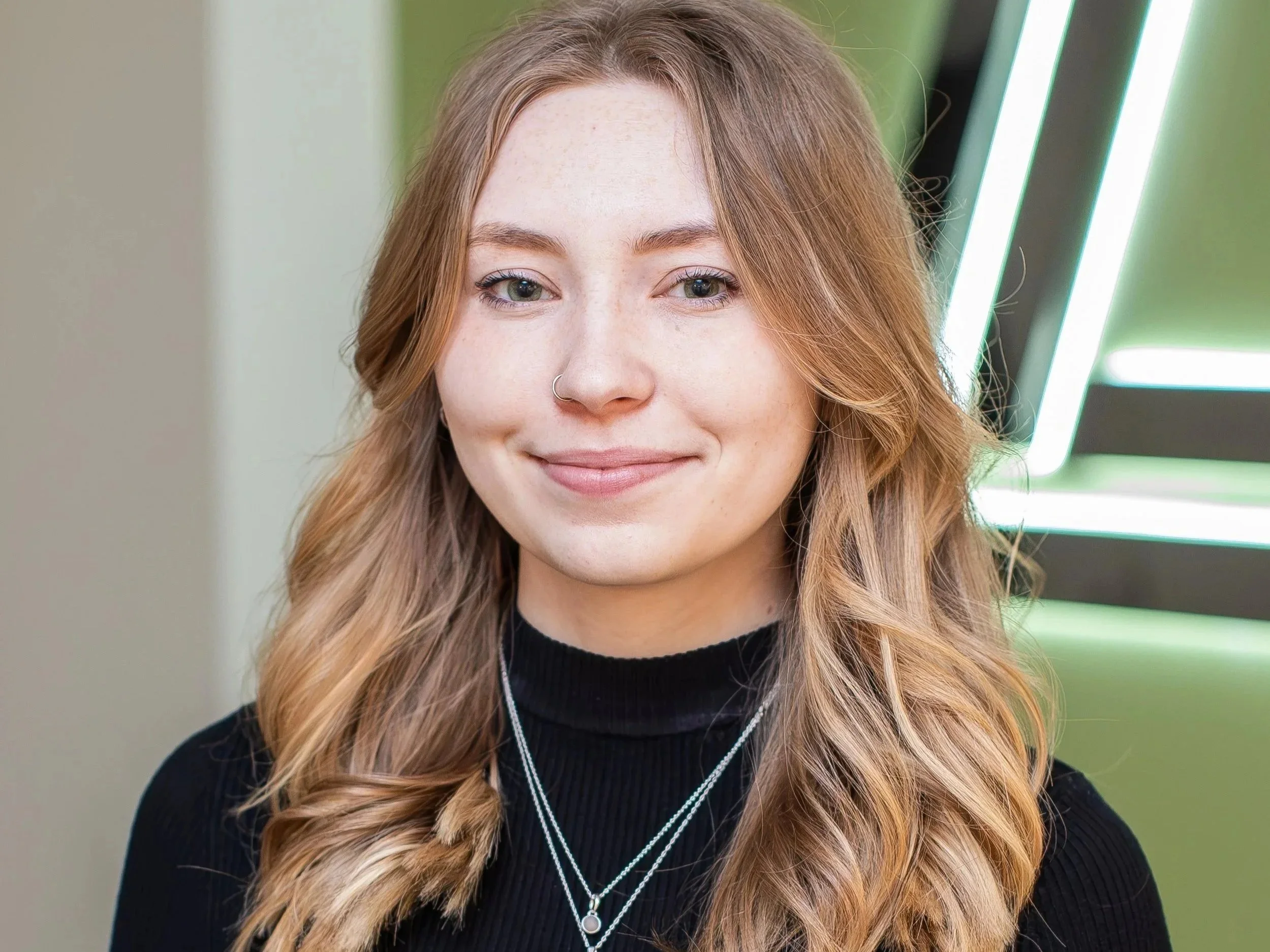 A young woman with blonde wavy hair, wearing a black turtleneck and layered necklaces, smiling indoors with a green wall and decorative lights in the background.