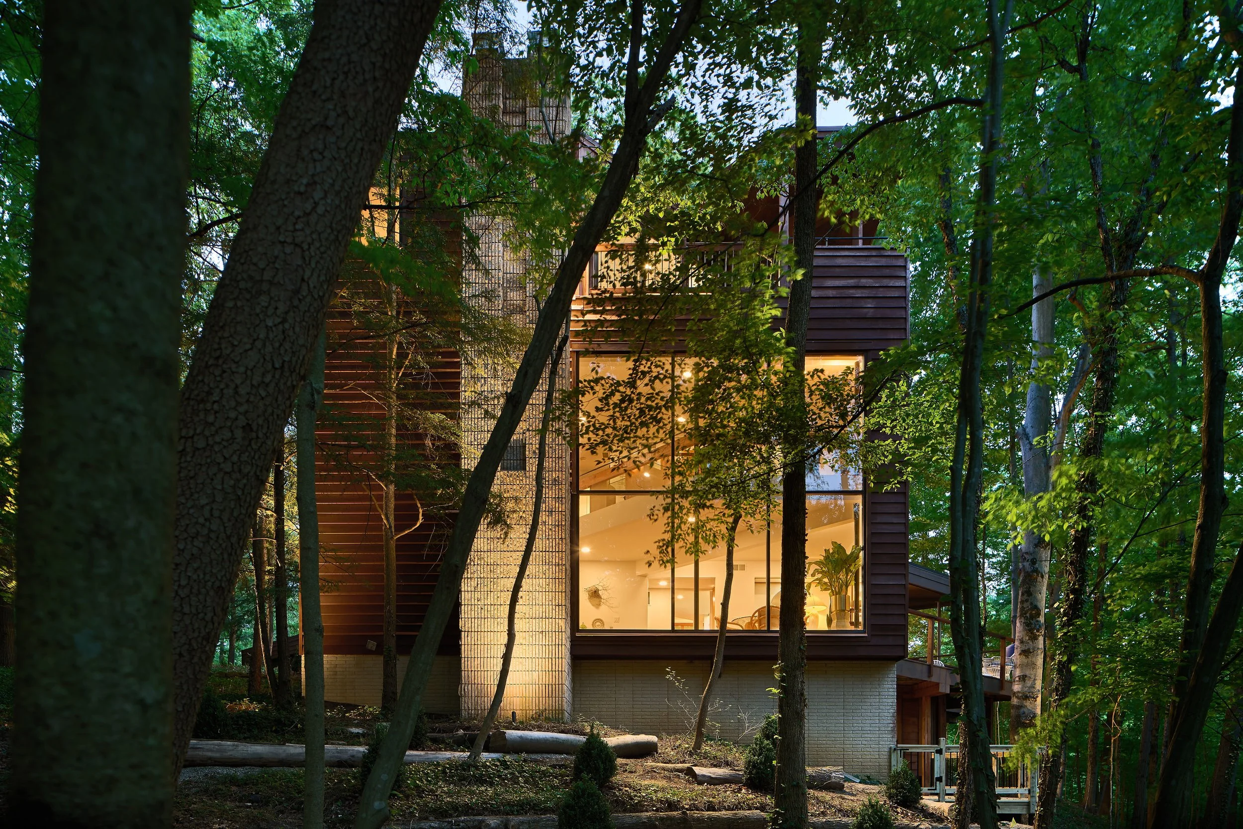 Modern house with large glass windows surrounded by trees, illuminated from inside, with a backyard visible through the glass.