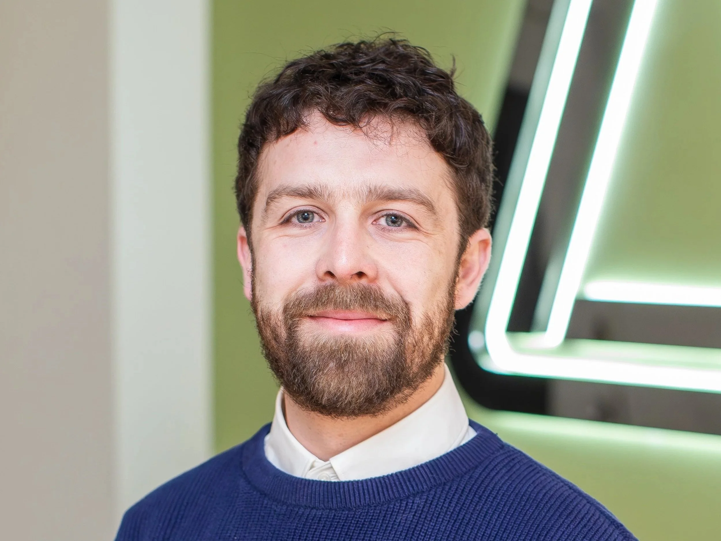 Close-up of a man with short dark hair, beard, and mustache, wearing a blue sweater over a white collared shirt, standing indoors with a green and black geometric background.