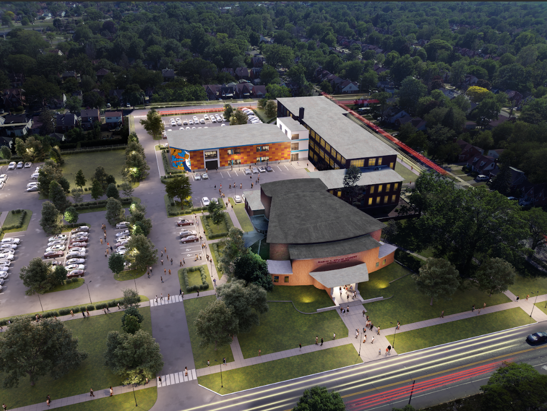 An aerial view of a modern school campus surrounded by trees and parking lots, with several people walking on sidewalks and the front entrance of the school.