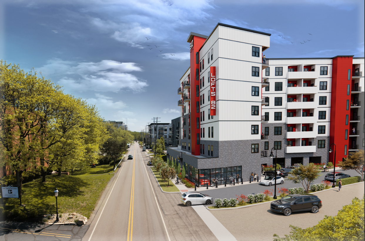 A modern multi-story apartment building with a red and white exterior, labeled 'Lofts 802,' situated on a city street with parked cars, pedestrians, trees, and a clear sky.