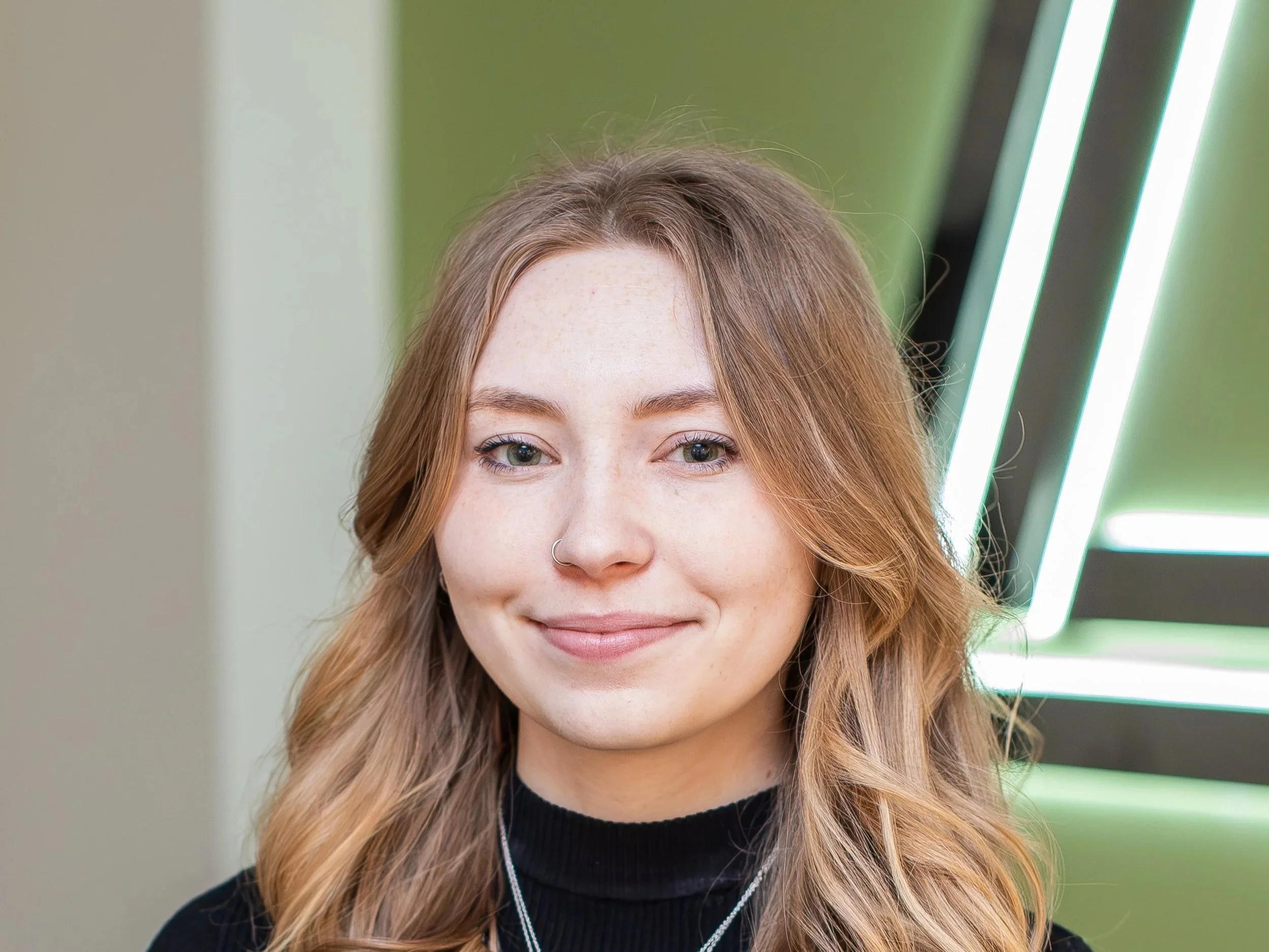 Portrait of a young woman with long wavy light brown hair, fair skin, a nose piercing, in front of a green and black patterned background