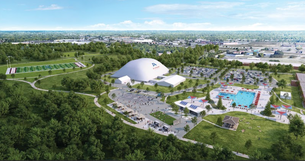 Aerial view of a park with a large domed arena, a swimming pool, a parking lot, and green fields, set in a suburban area.