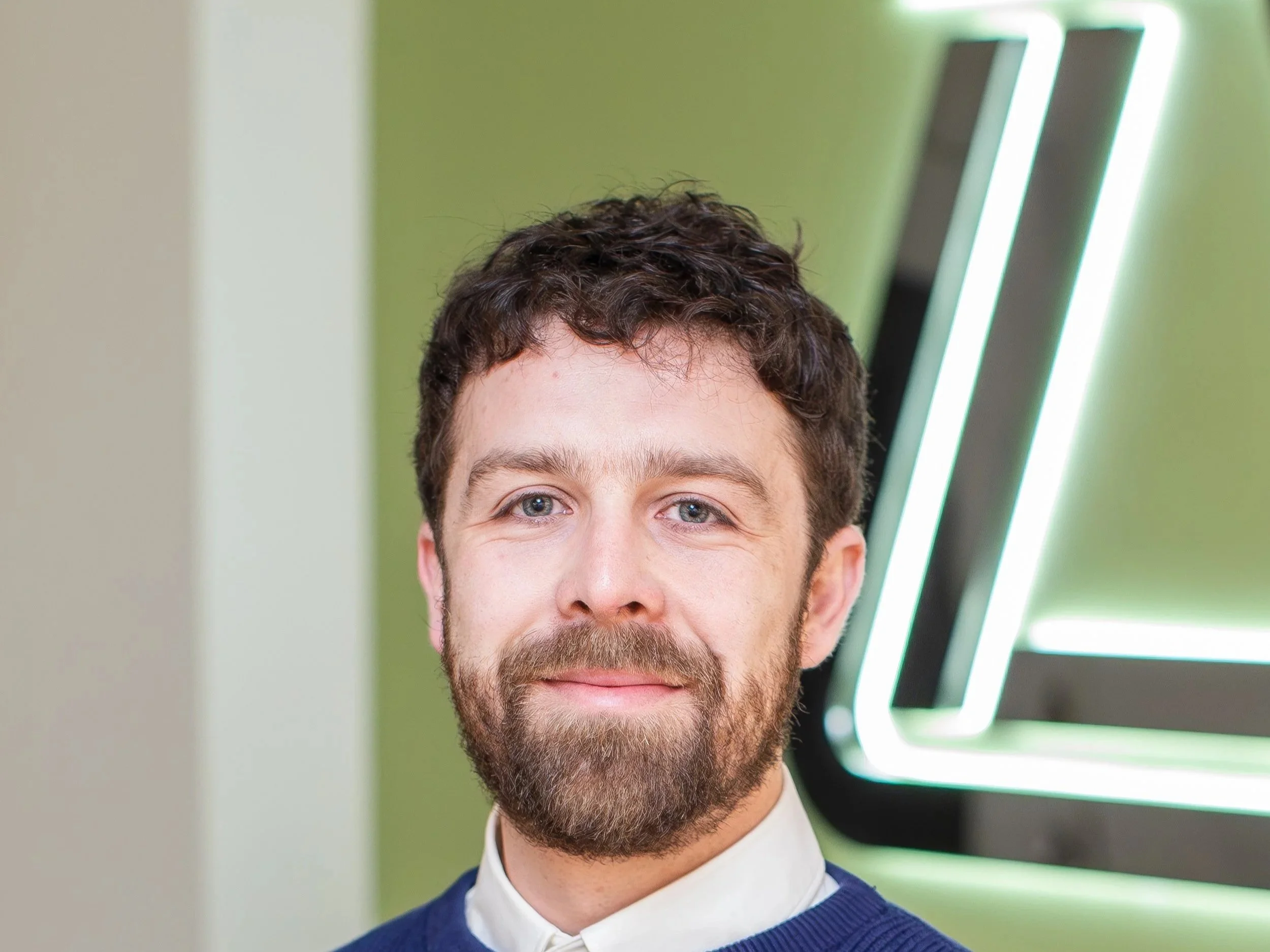 A man with dark curly hair and a beard, wearing a white collared shirt and a dark blue sweater, standing indoors with green walls and illuminated modern art or signage in the background.