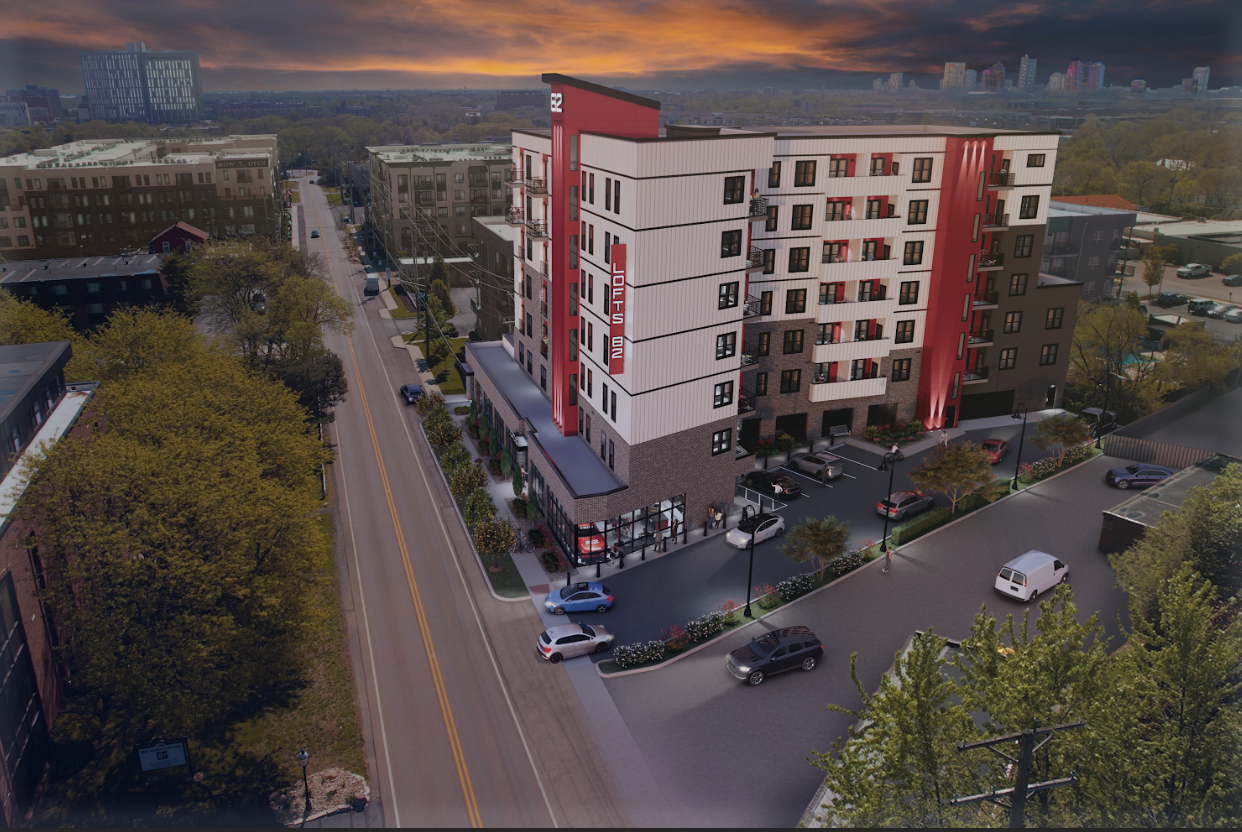 An aerial view of a modern hotel named 'Lofters,' a five-story building with a parking lot in front, located at the corner of two streets in an urban area during sunset.