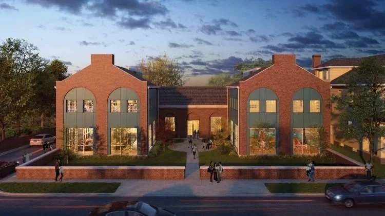 A modern brick building with large windows and two arched sections, surrounded by a lawn and pathway, with trees and a street in the foreground on a partly cloudy evening.