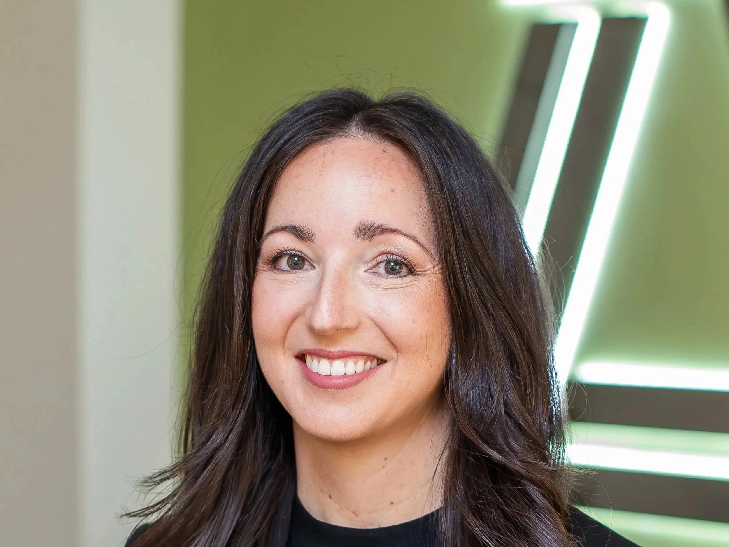 A woman with long dark hair smiling in front of a modern, abstract light fixture on a green wall.