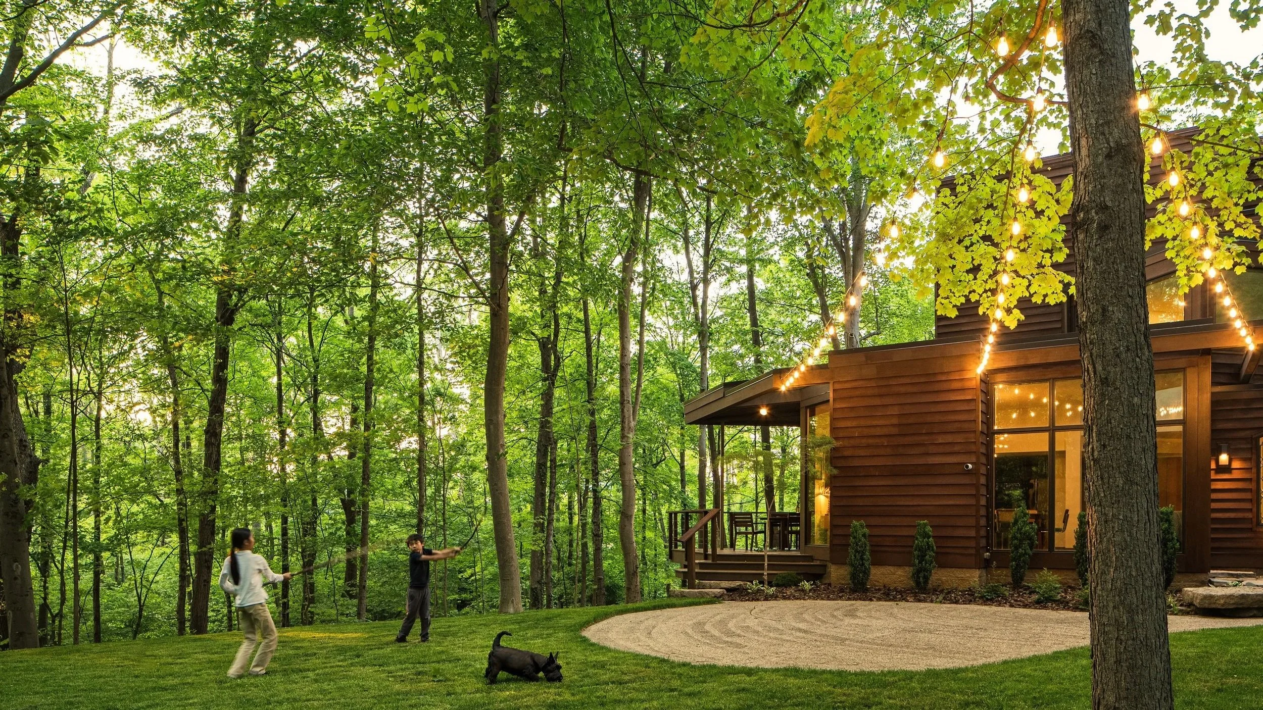 Children playing with a dog in front of a modern wooden house with string lights, surrounded by a lush green forest.