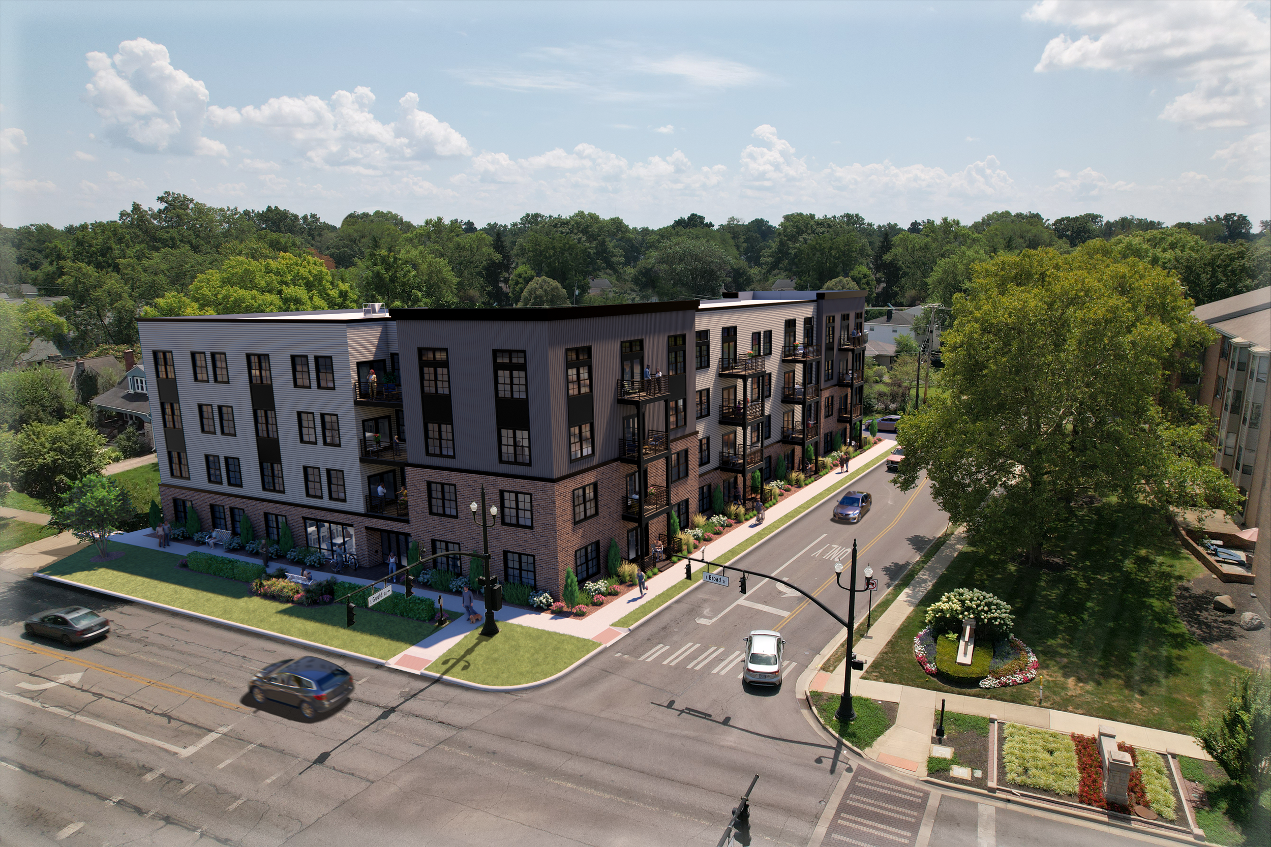 A modern four-story apartment building with balconies, located on a street corner with a manicured lawn, sidewalk, and street trees, in a neighborhood with other residential buildings and a wooded area in the background.
