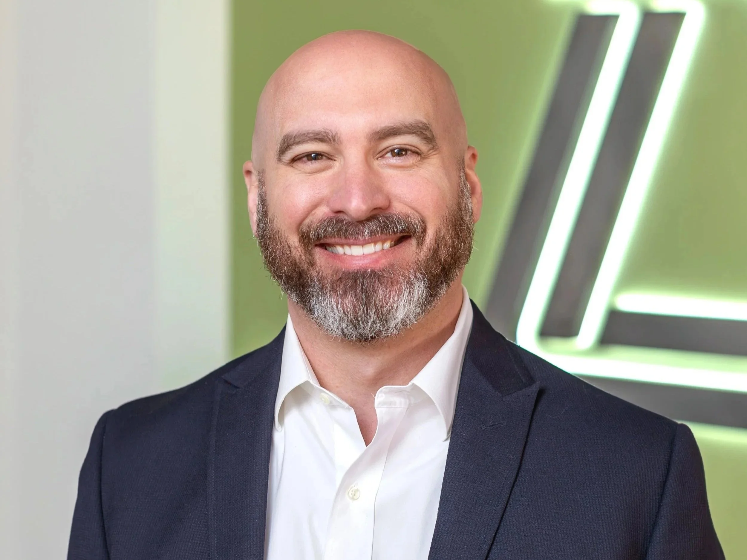 A smiling bald man with a beard wearing a white dress shirt and a navy blazer, standing indoors with green and neon-lit geometric wall art in the background.