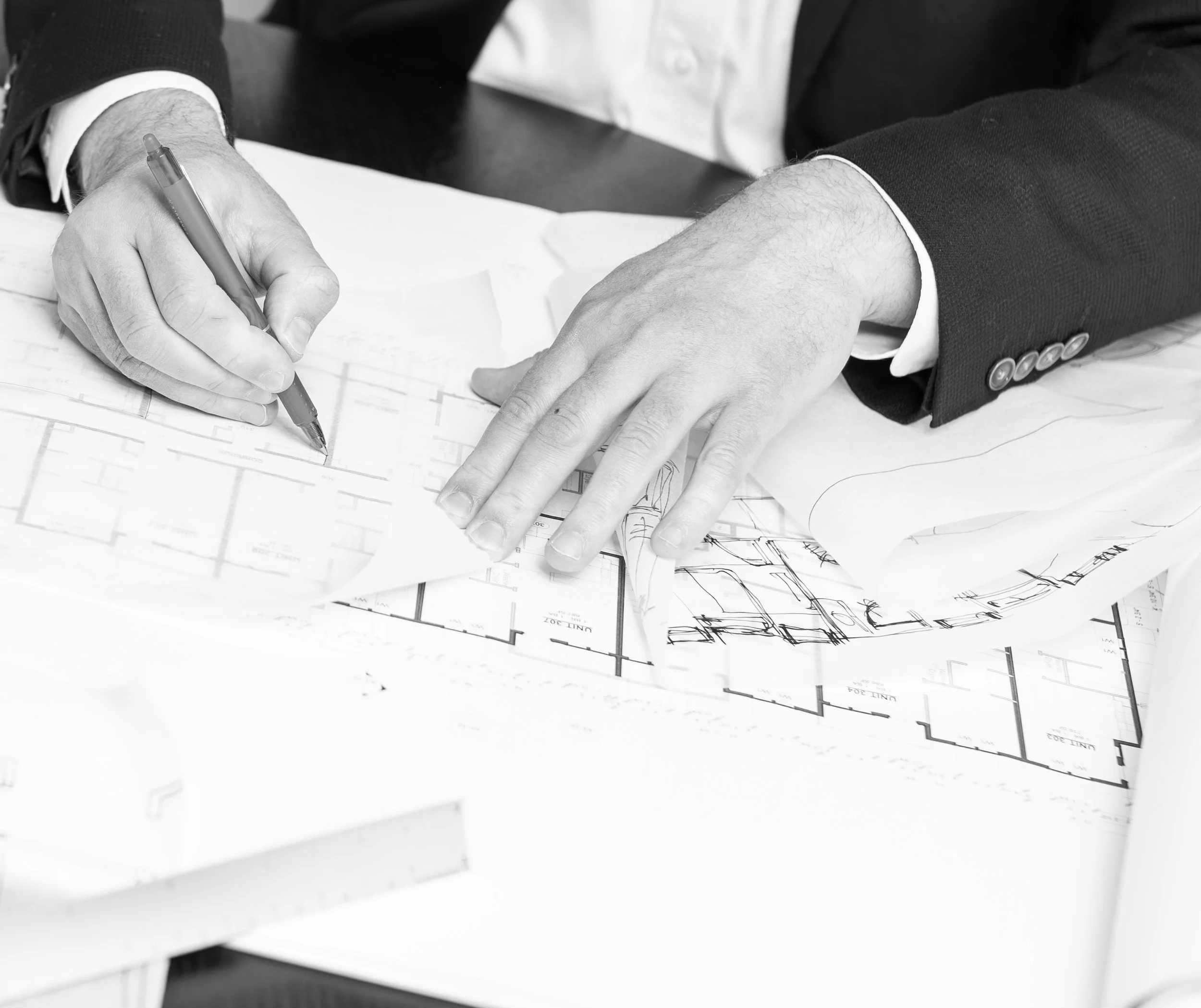 A man in a suit using a pen to draw on architectural blueprints on a table, with detailed floor plans visible.