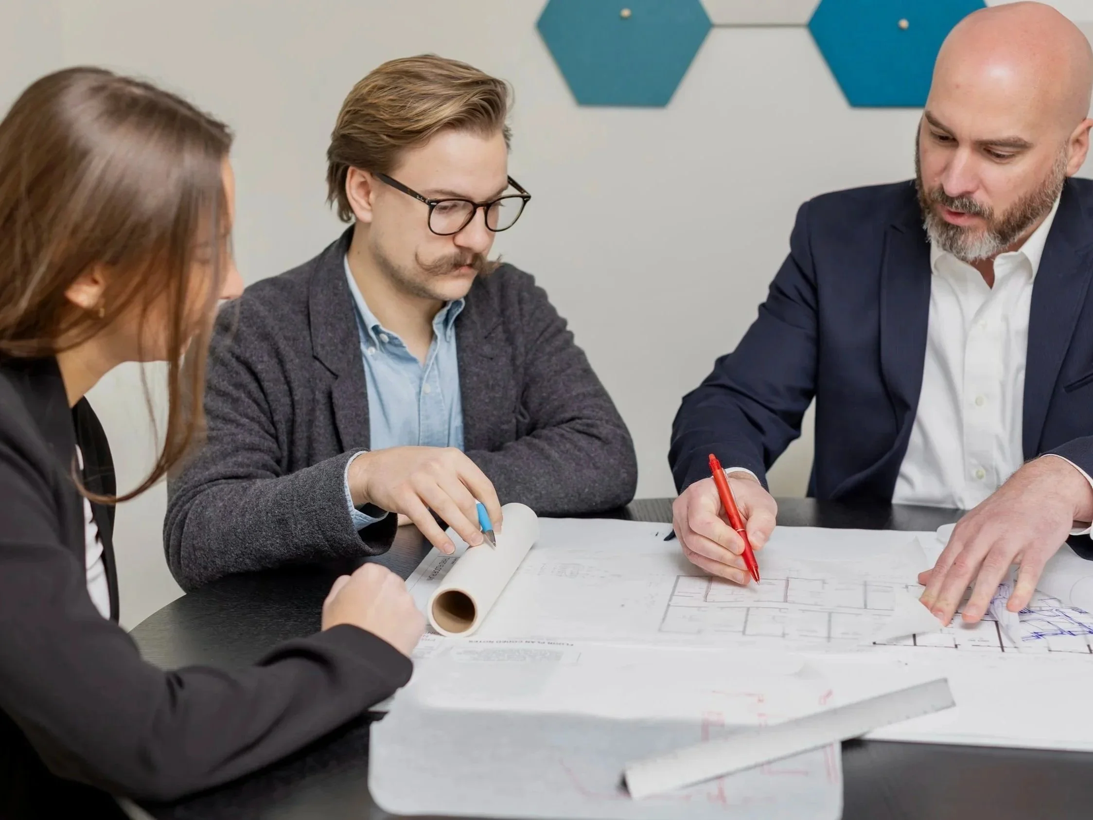 Three professionals at a table reviewing architectural blueprints, with one using a ruler and another pointing with a red pen.