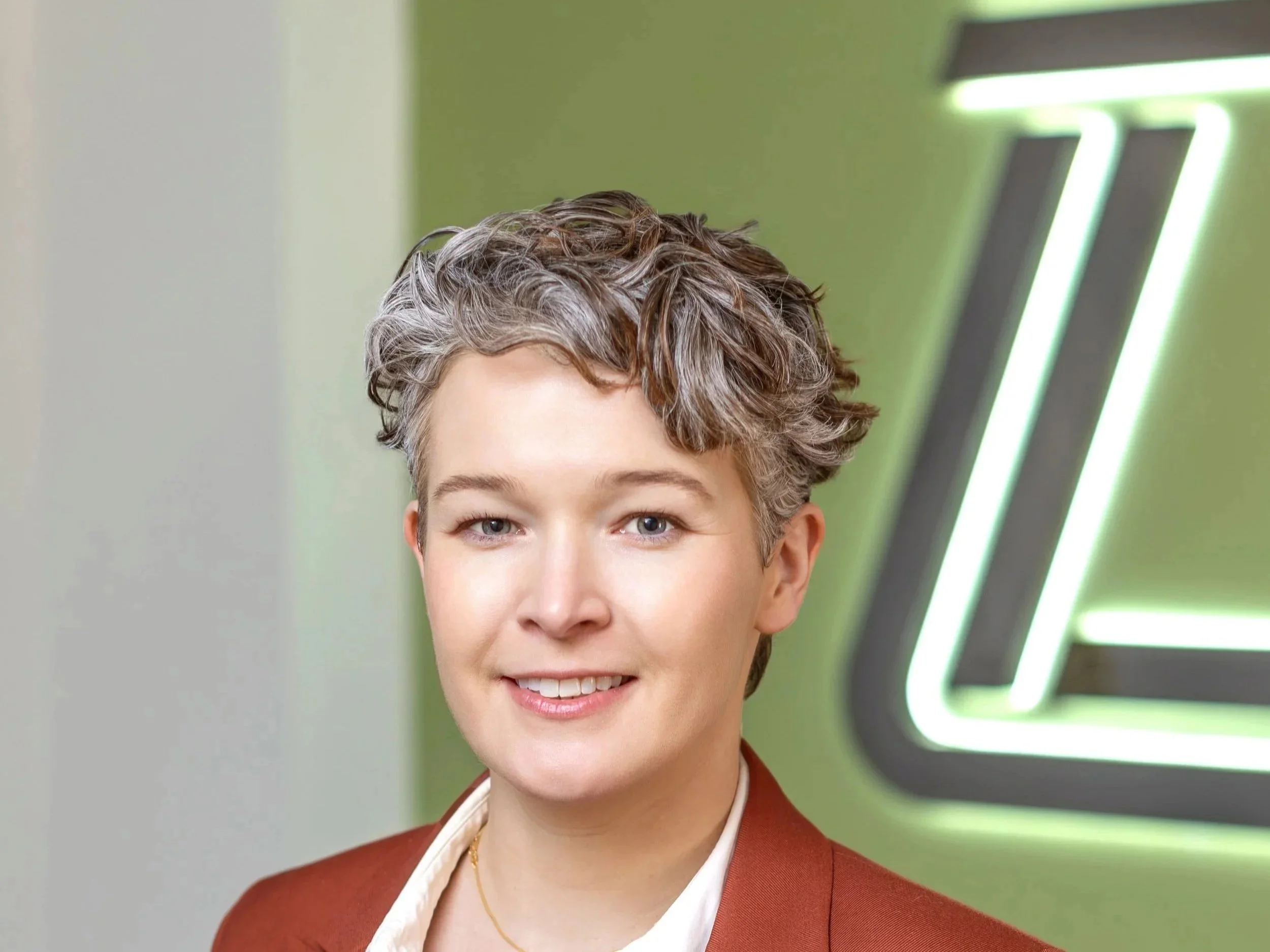 A woman with short, wavy gray hair and fair skin smiling in front of a green wall with neon lights.