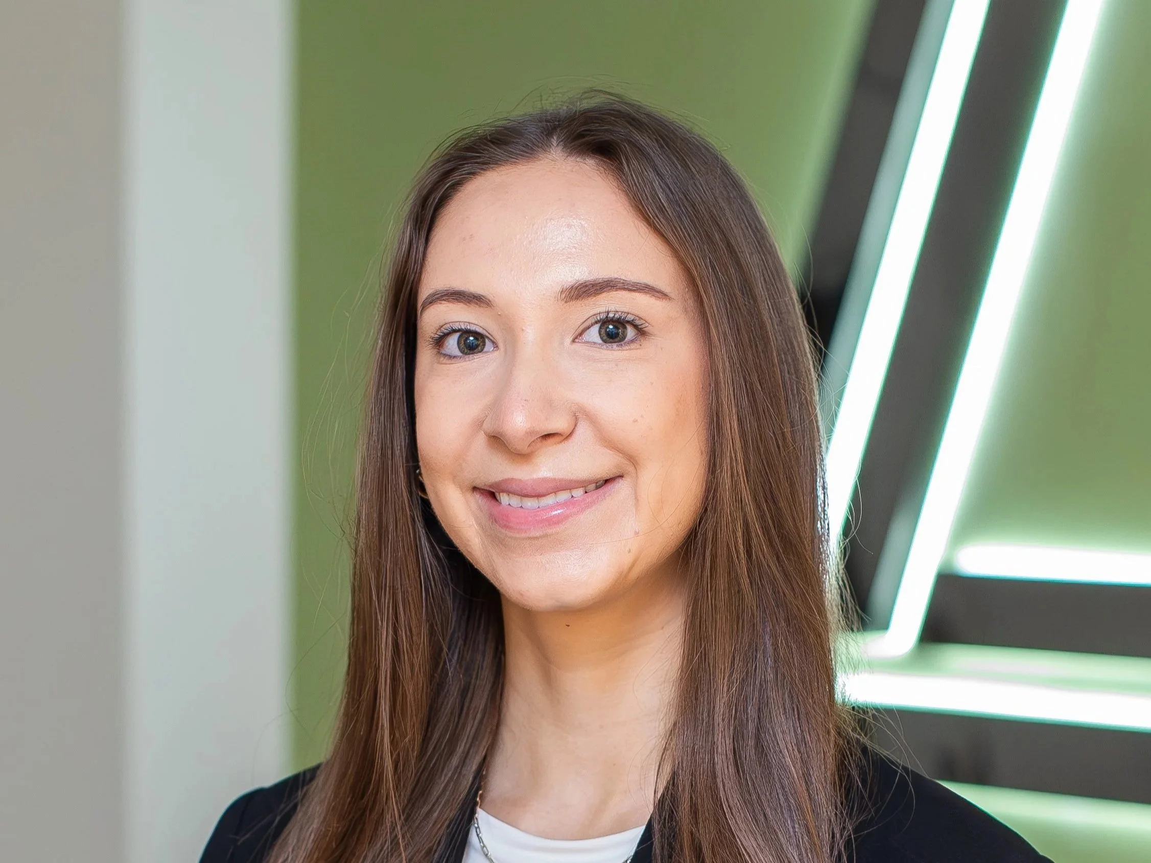 A smiling woman with long brown hair, wearing a black blazer over a white top, standing in front of a modern green wall with illuminated design elements.