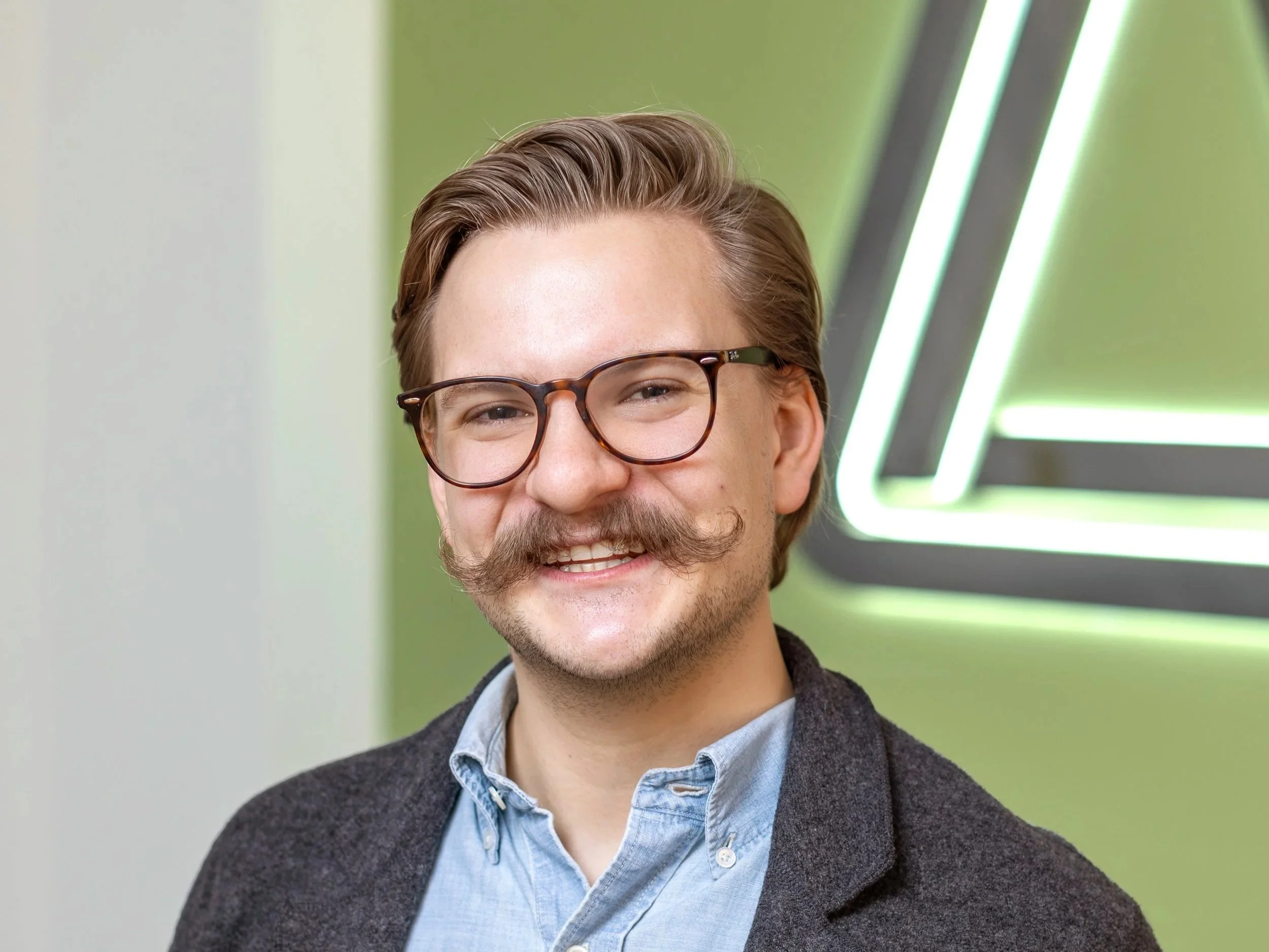 A smiling man with glasses, a mustache, and a beard, wearing a light blue shirt and a dark blazer, standing in front of a green wall with a neon sign.