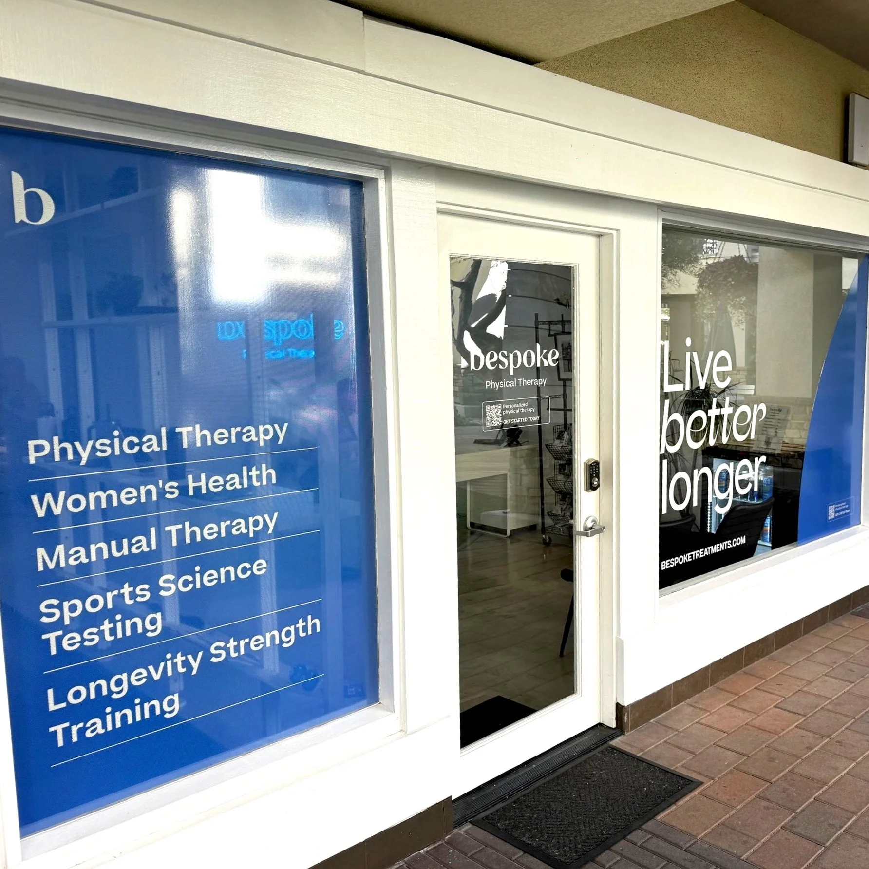 Storefront window displaying services like physical therapy, women's health, manual therapy, sports science testing, and longevity training. The entry door has a sign saying 'Bespoke' and the phrase 'Live better longer' is on the right window.