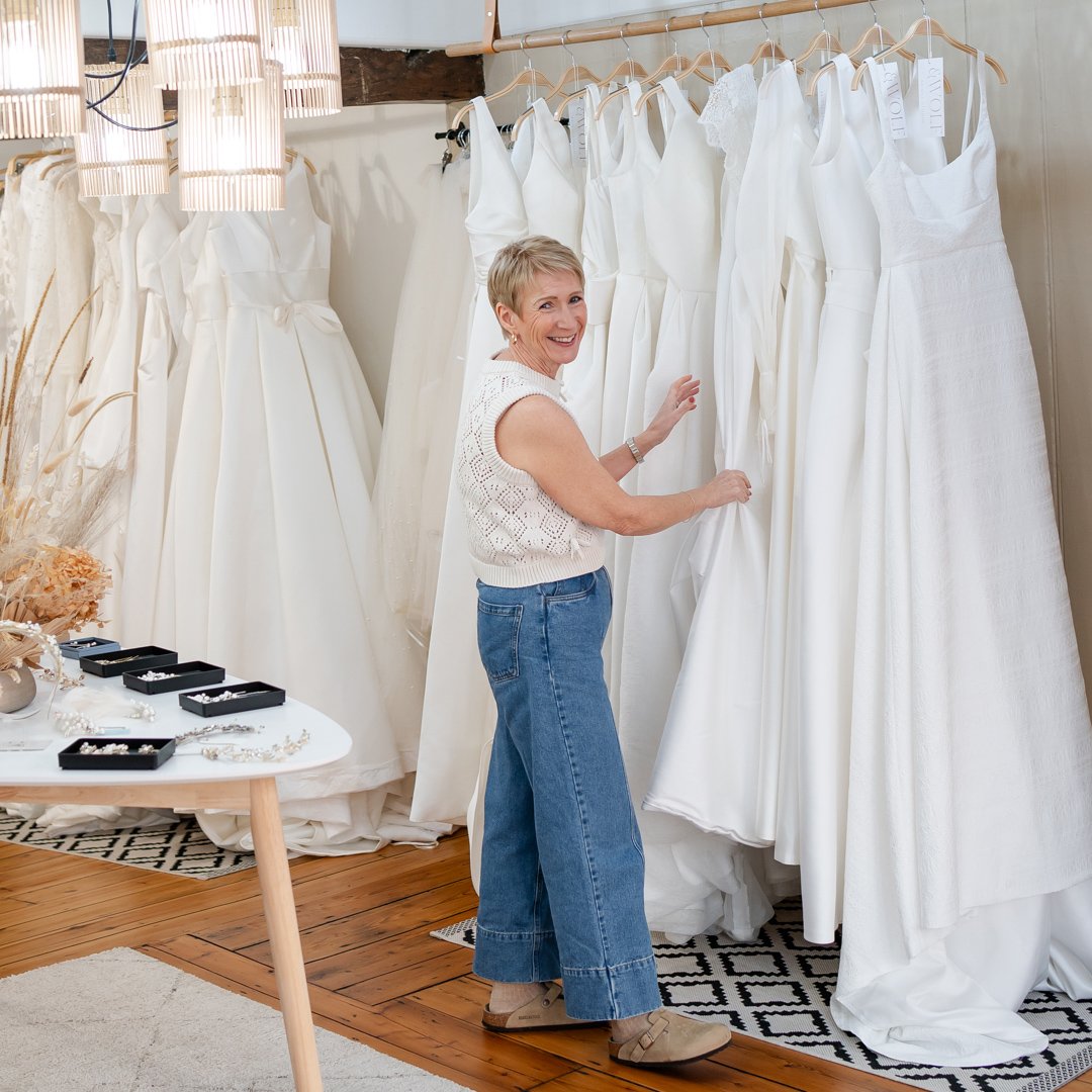 A smiling woman looking at white wedding dresses hanging on a rack in a bridal boutique.