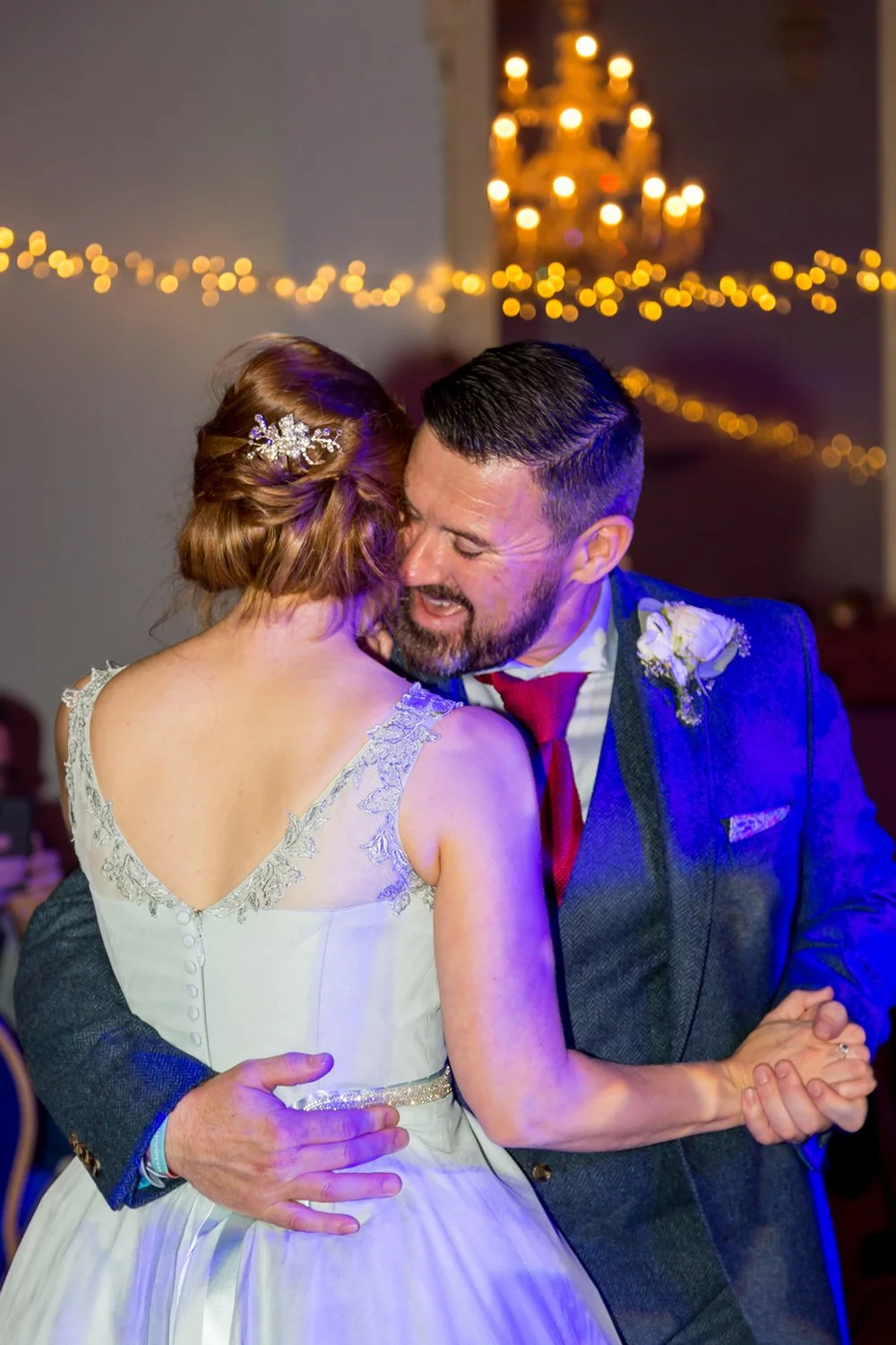 A couple dancing closely at a wedding reception, with fairy lights and a chandelier in the background.