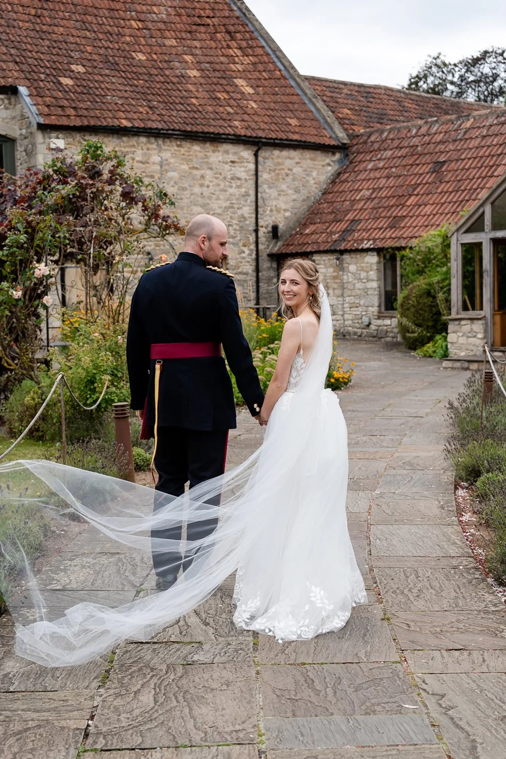 A bride and groom holding hands, smiling and looking at each other outside in a garden, with a rustic stone building and garden flowers in the background.