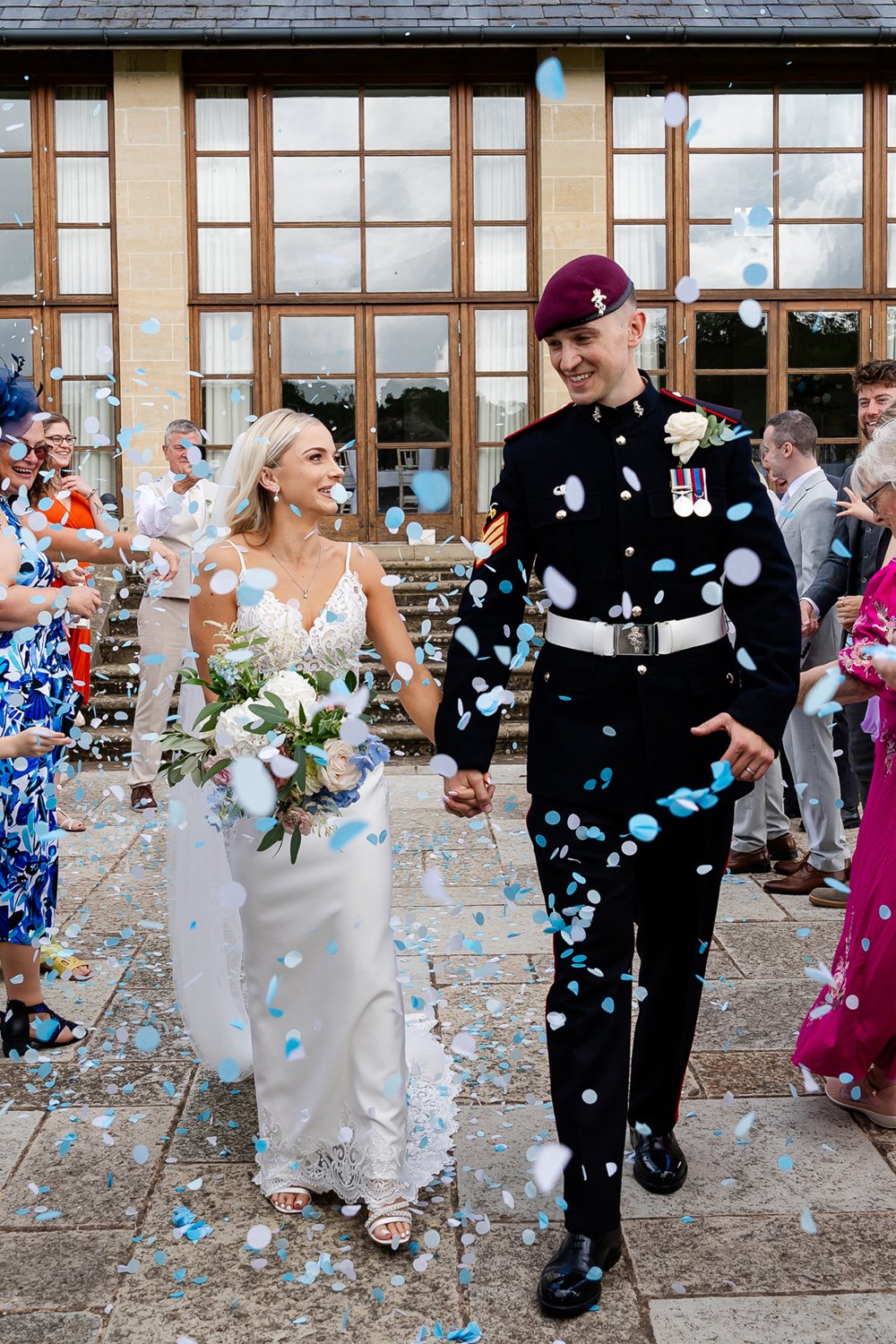 A bride and groom holding hands and walking through falling blue and white confetti outside a building with large windows, surrounded by smiling guests during their wedding celebration.