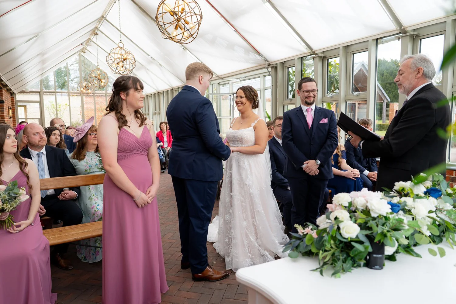 A wedding ceremony taking place inside a bright greenhouse with large windows and white walls. The bride and groom are holding hands and facing each other, exchanging vows, with a officiant reading from a black folder. The bride is wearing a white lace wedding dress, and the groom is dressed in a navy blue suit. Bridesmaids and groomsmen are seated and standing nearby, with some women wearing pink dresses. Guests are seated in the background, watching the ceremony.