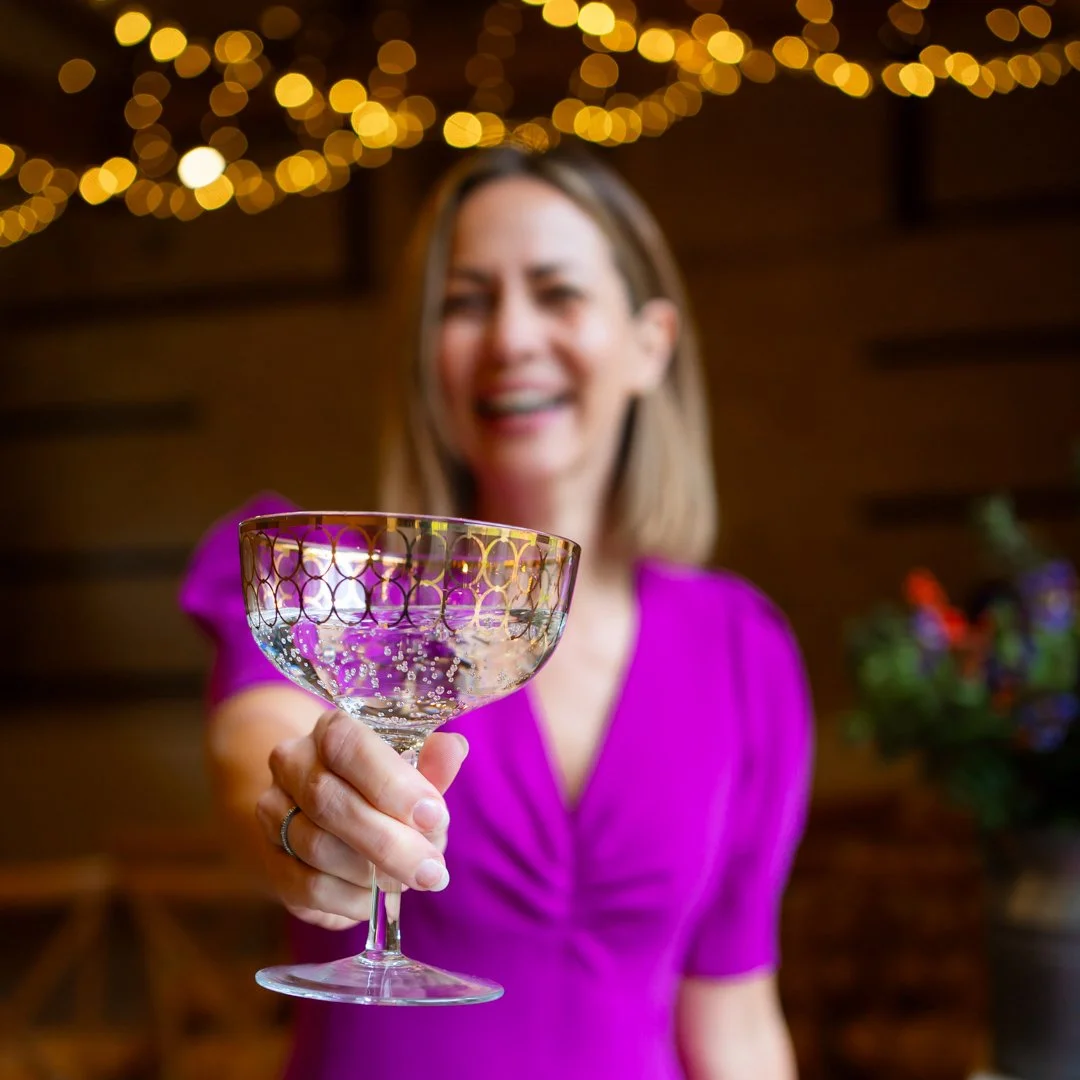 A woman in a magenta dress smiling while holding a colorful cocktail glass at a celebration with warm lighting and blurred background.
