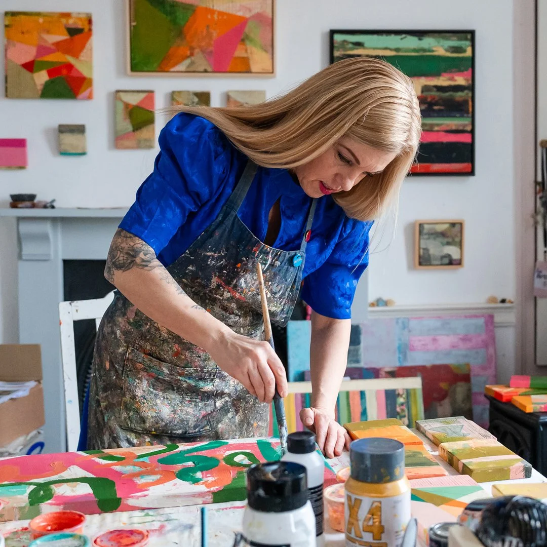 A woman with long blonde hair painting abstract artwork in a colorful art studio surrounded by paintings and art supplies.