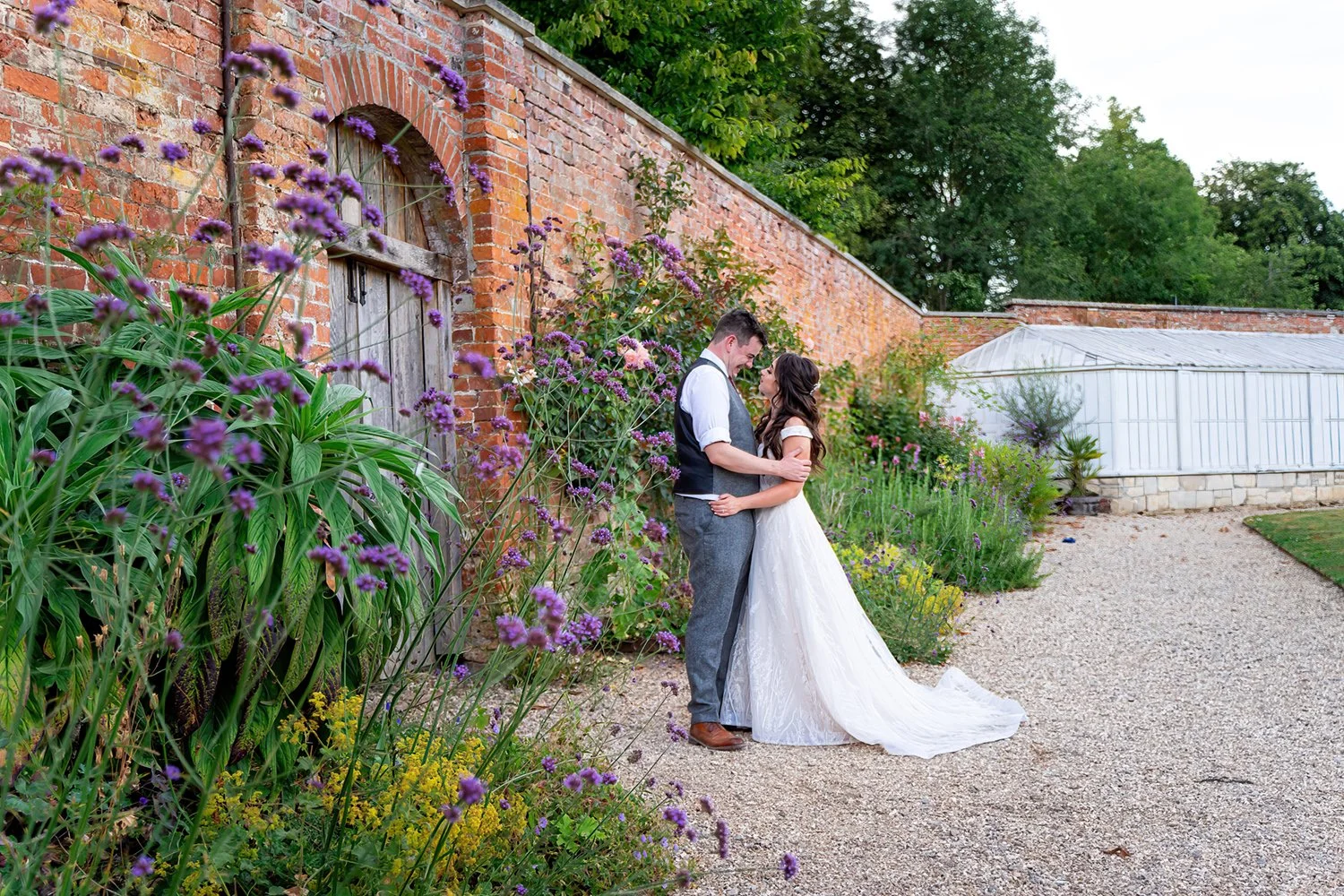 A bride and groom standing close together by a brick wall with purple flowers, outdoors during daytime.