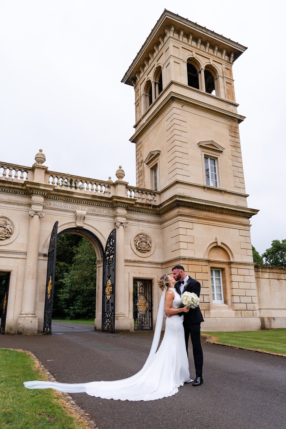 Bride and groom in wedding attire sharing a kiss outside the Golden Gates of Bowood in Wiltshire.