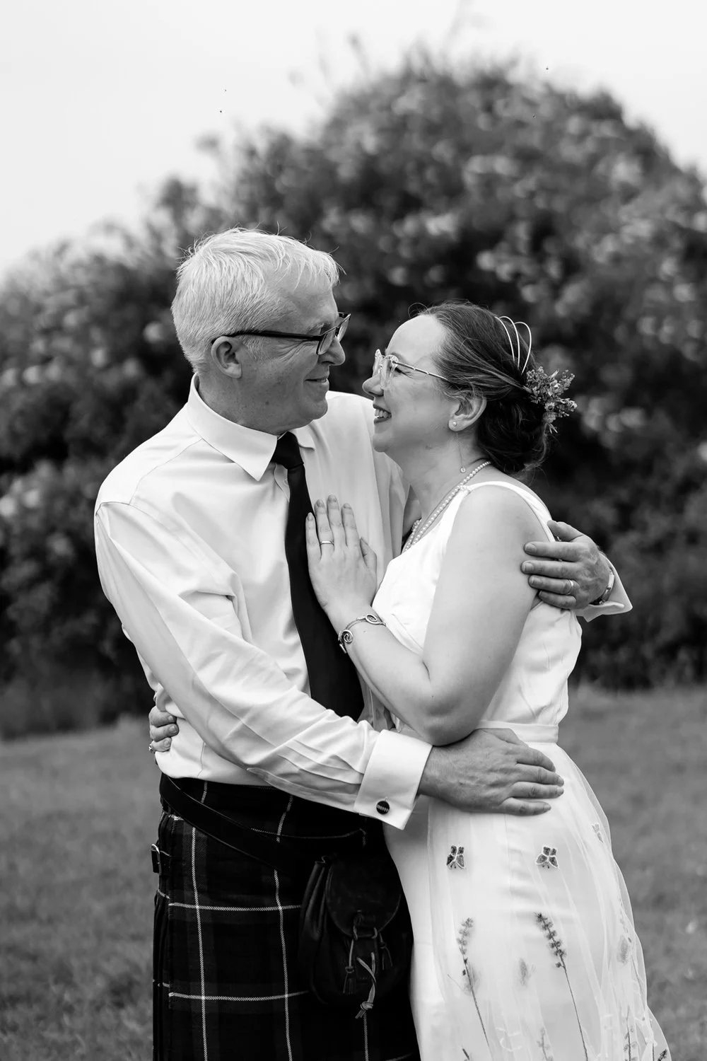 A happy couple in wedding attire embracing outdoors, smiling at each other.