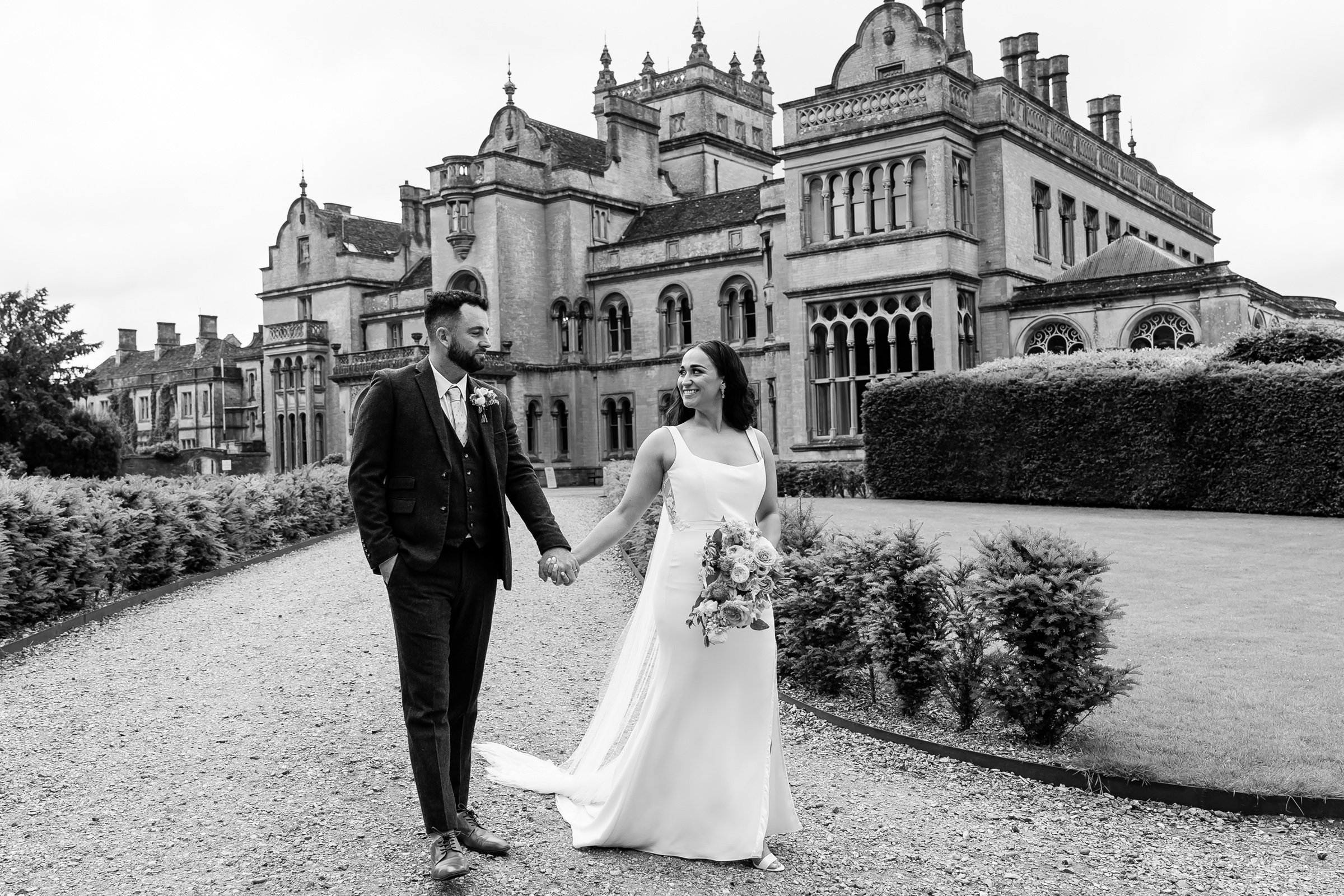A black-and-white photo of a newlywed couple walking hand in hand outdoors in front of a castle-like building. The bride is wearing a white wedding dress and holding a bouquet of flowers, and the groom is wearing a dark suit with a boutonnière.