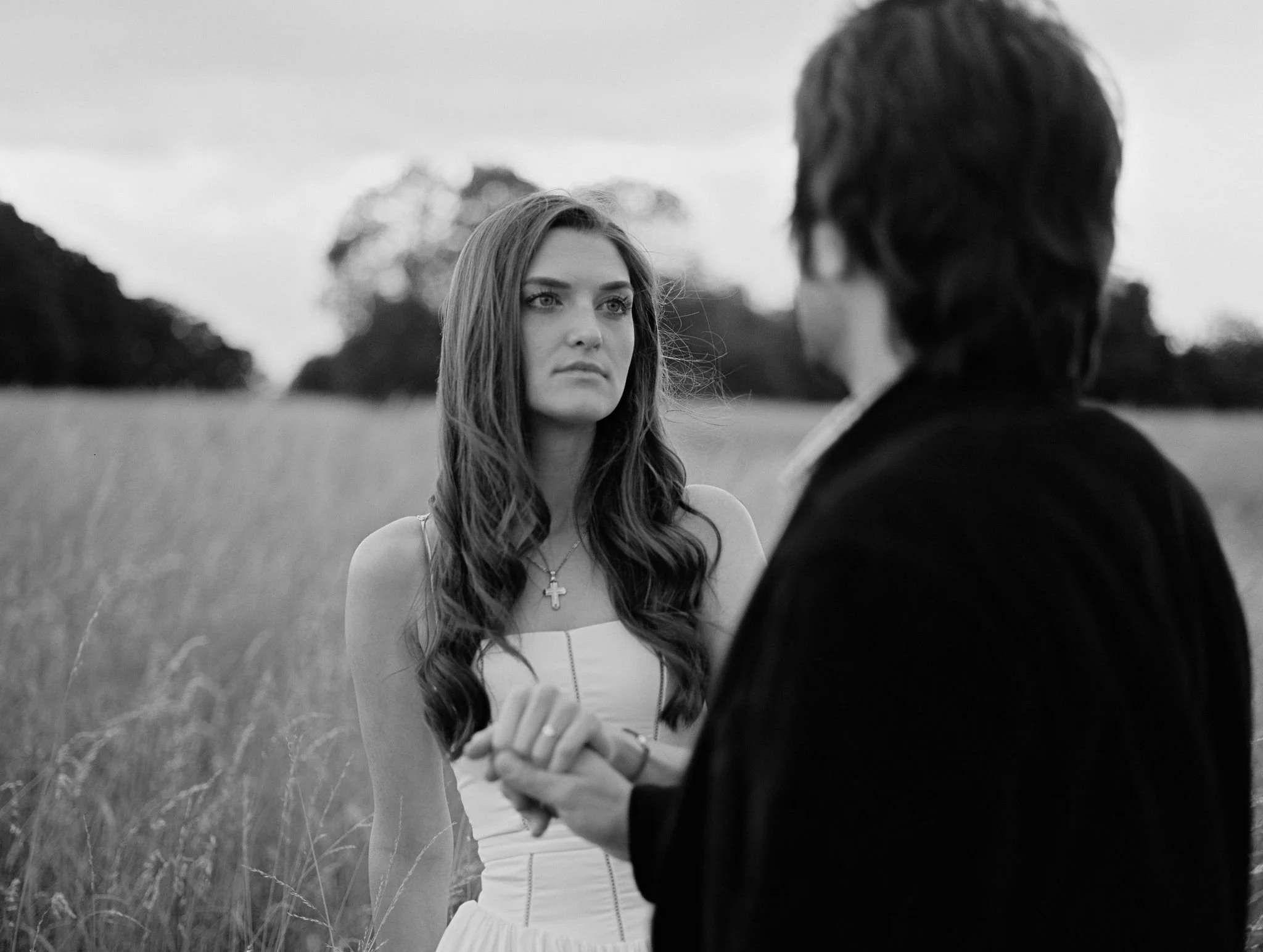 A black-and-white photo of a woman with long hair in a white dress holding hands with a man in a dark jacket in an outdoor field.