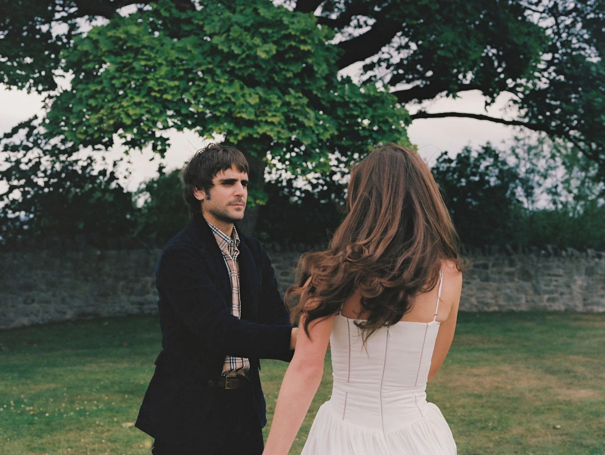 A man and a woman facing each other outdoors with a large tree and stone wall in the background.