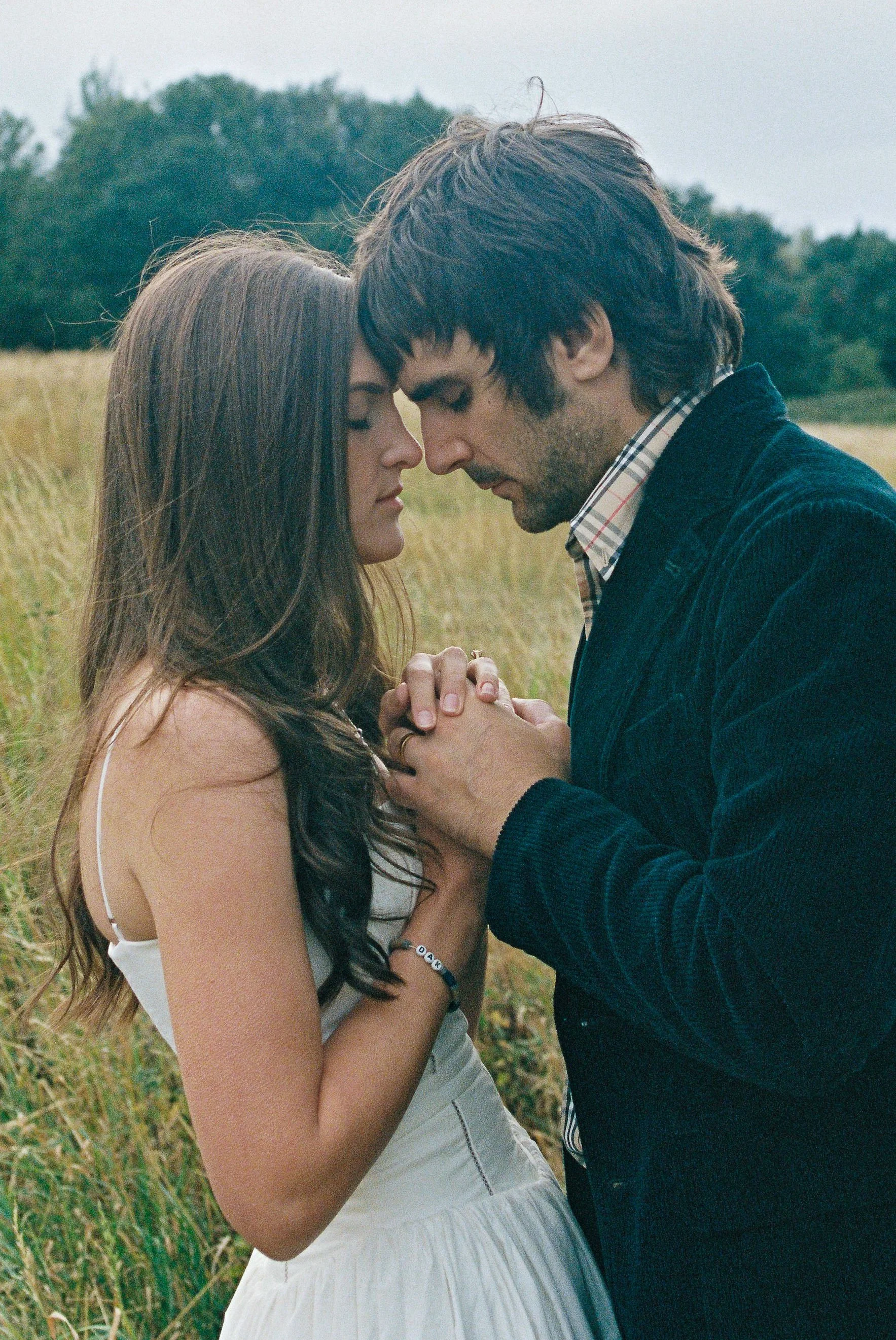 A young couple standing close together in a field, with their foreheads touching and hands clasped, sharing an intimate moment.