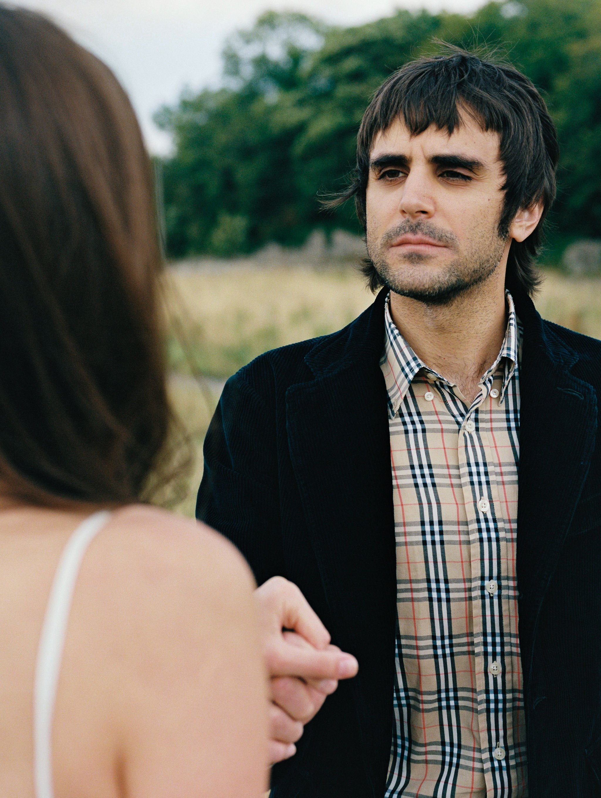 A man with dark hair and a beard standing outdoors in front of trees, wearing a plaid shirt and a black jacket, looking serious. Part of a woman's shoulder and arm is visible in the foreground.
