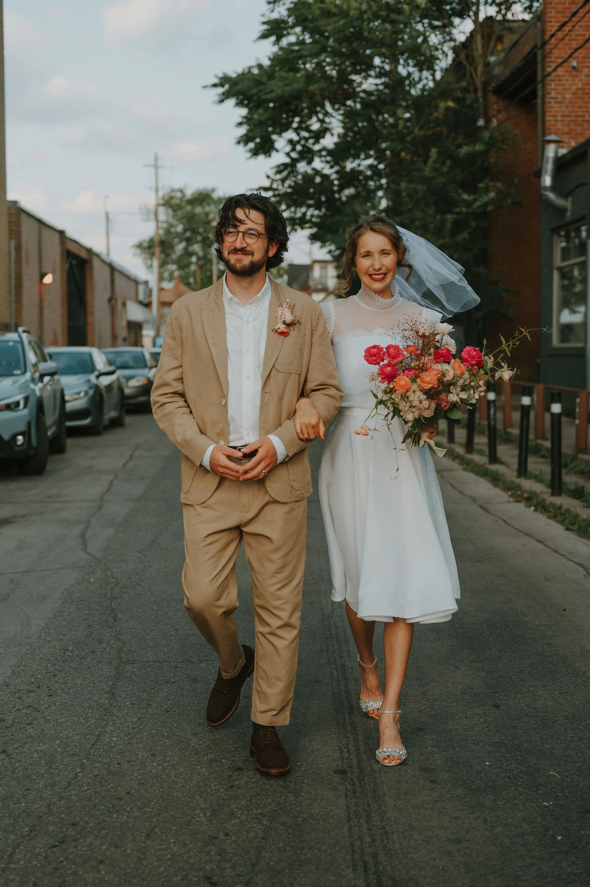 A bride and groom walking together on a city street during their wedding, with the bride holding a large bouquet of pink and orange flowers and the groom wearing a beige suit.
