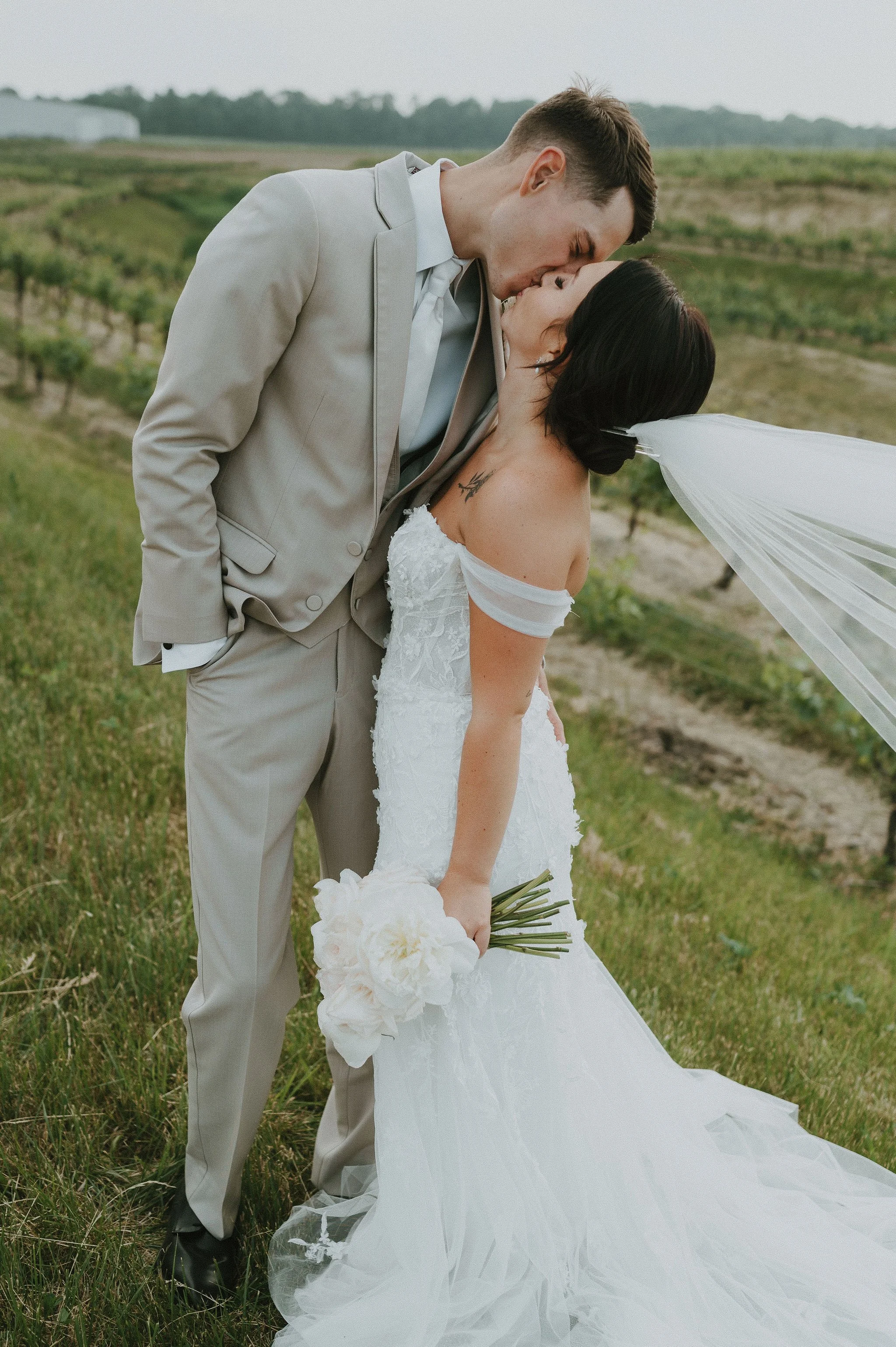 A newlywed couple kissing in a vineyard, the groom leaning over the bride, who holds a bouquet of white flowers, the bride's veil flowing in the wind, both dressed in wedding attire.